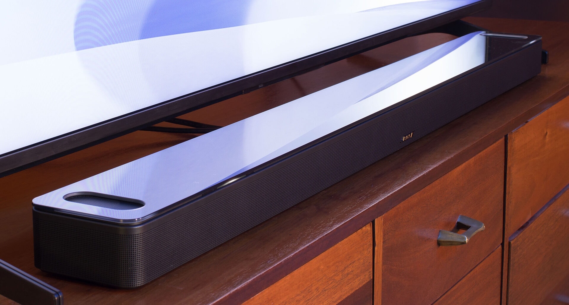 Nahaufnahme einer schwarzen Soundbar mit Glasoberfläche vor einem TV-Gerät Nahaufnahme einer schwarzen Soundbar mit Glasoberfläche vor einem TV-Gerät