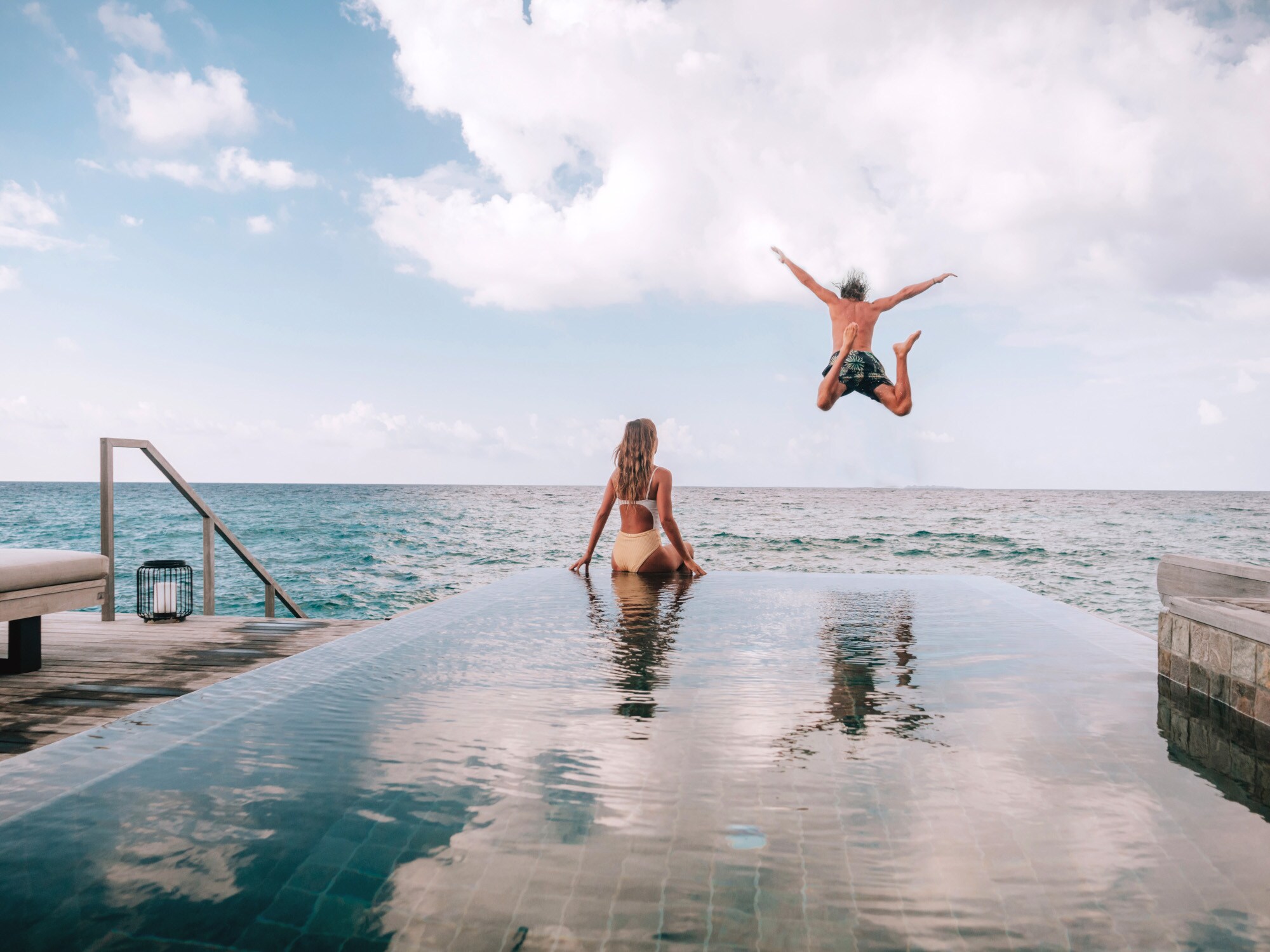 Eine Frau sitzt am Rande eines Pools am Meer, während ein Mann ins Meer springt Eine Frau sitzt am Rande eines Pools am Meer, während ein Mann ins Meer springt