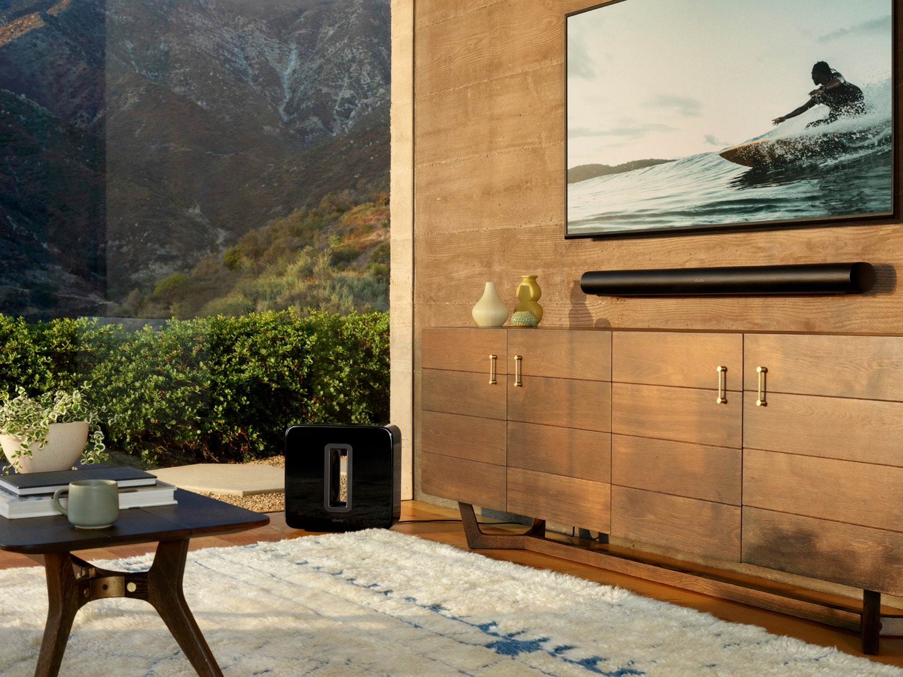 Ein modernes Wohnzimmer mit Panoramafenster sowie einem TV-Gerät und einer Soundbar an der Wand, daneben ein Subwoofer auf dem Boden Ein modernes Wohnzimmer mit Panoramafenster sowie einem TV-Gerät und einer Soundbar an der Wand, daneben ein Subwoofer auf dem Boden