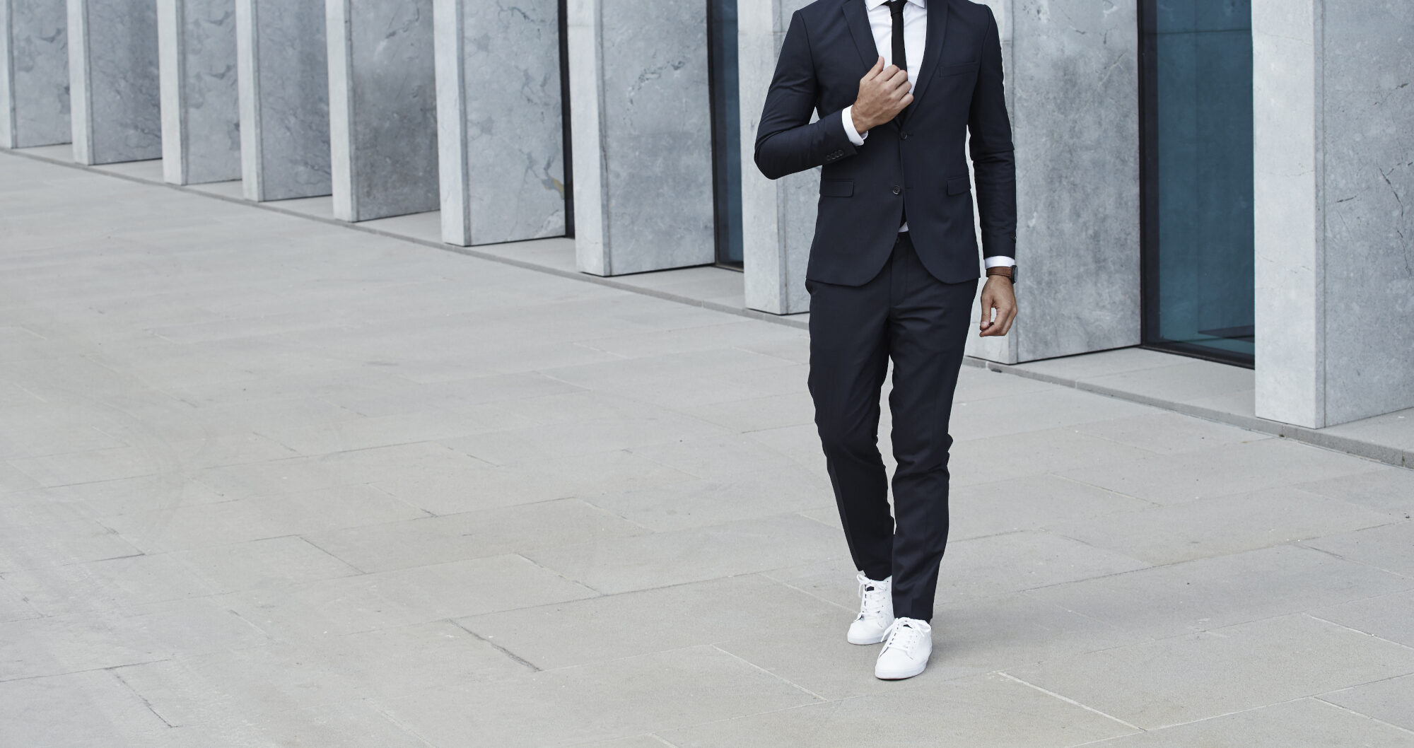 Ein Mann in einem dunklen Anzug und mit weißen Turnschuhen vor einem hellen Haus mit großen Fenstern Ein Mann in einem dunklen Anzug und mit weißen Turnschuhen vor einem hellen Haus mit großen Fenstern