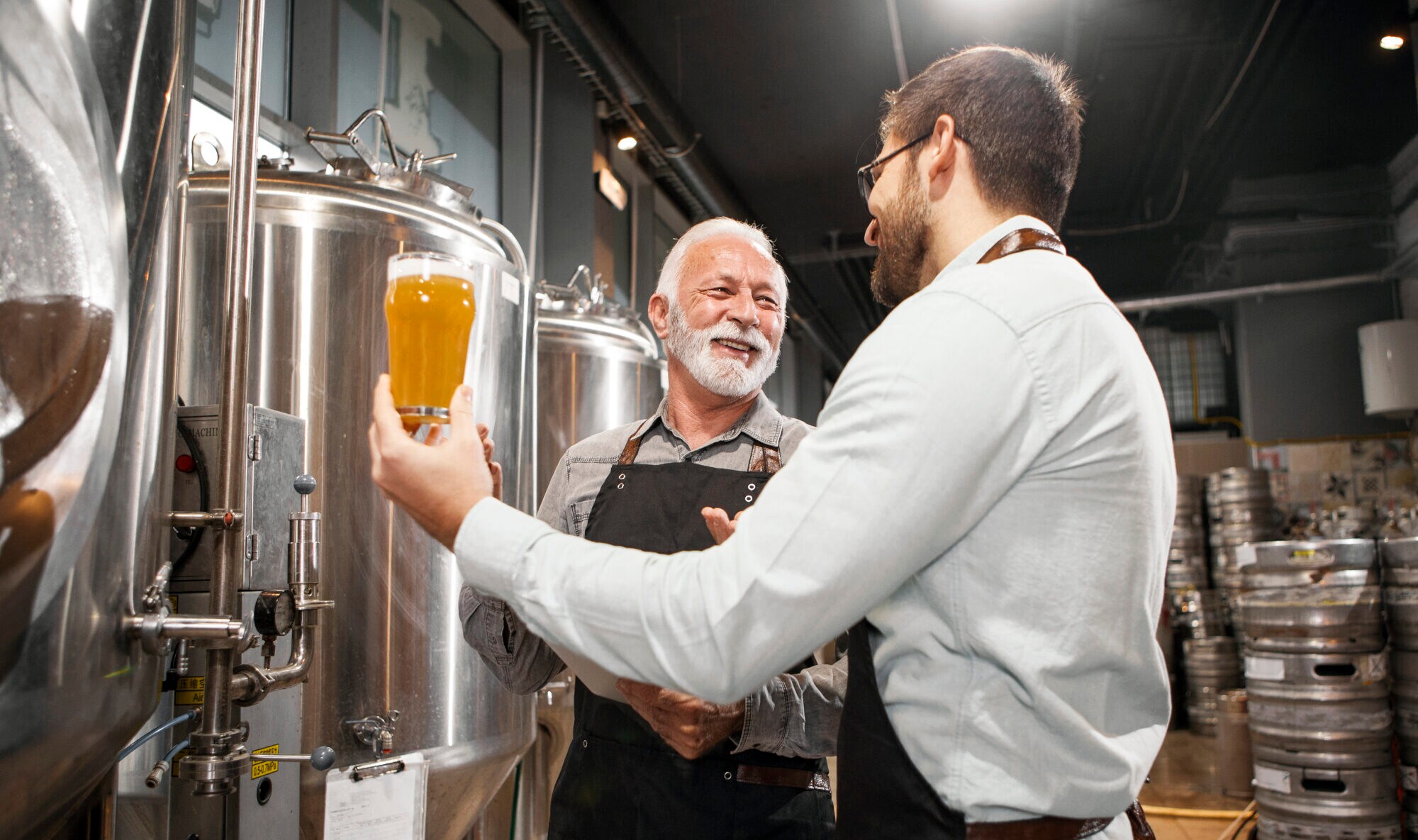 Zwei lächelnde Männer in einer Brauerei, von denen einer ein Glas Bier hochhält