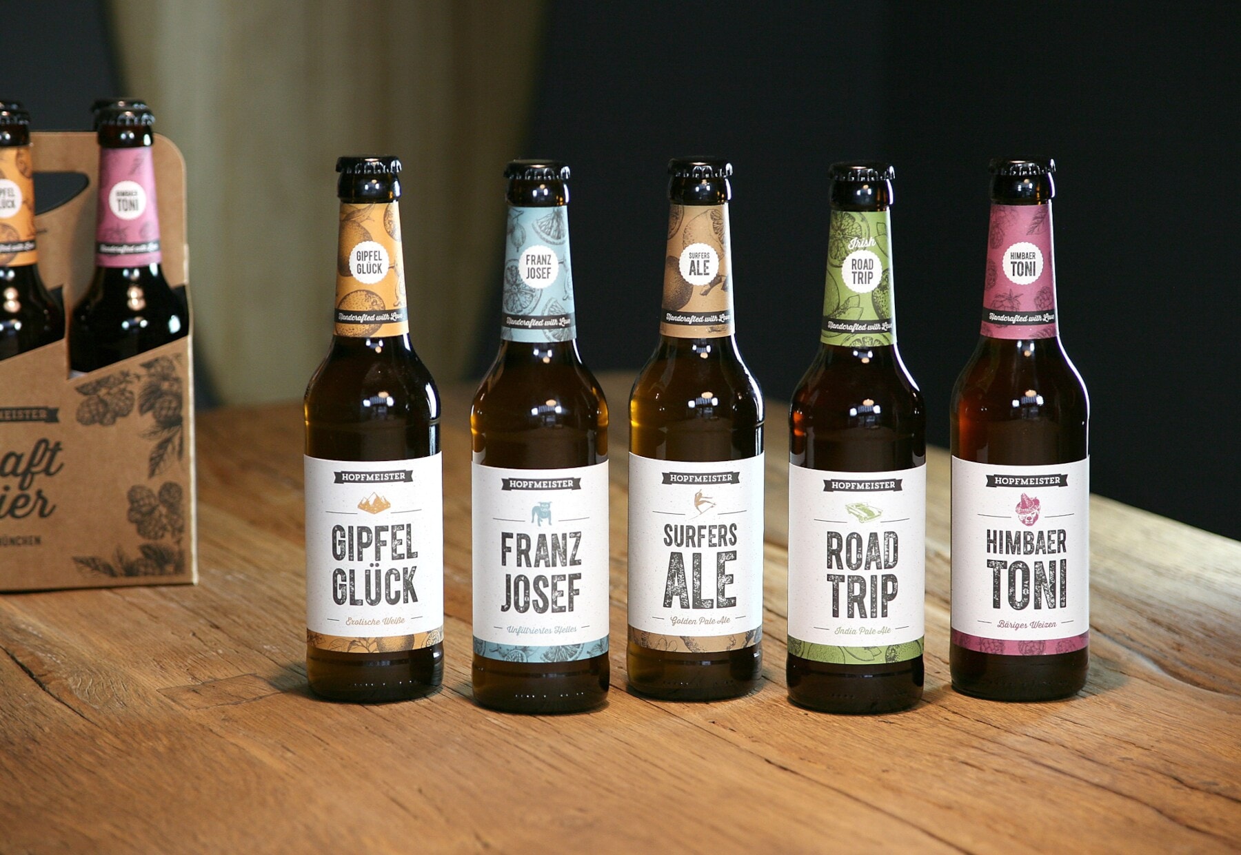 Mehrere Bierflaschen mit bunten Labels, teilweise in einem Karton, auf einem Holztisch Mehrere Bierflaschen mit bunten Labels, teilweise in einem Karton, auf einem Holztisch