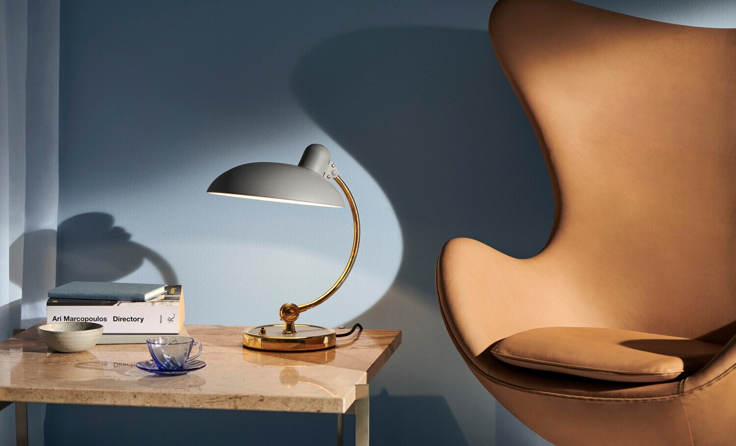 Neben einem Lesesessel steht die Tischleuchte Kaiser Idell auf einem Tisch. Neben einem Lesesessel steht die Tischleuchte Kaiser Idell auf einem Tisch.