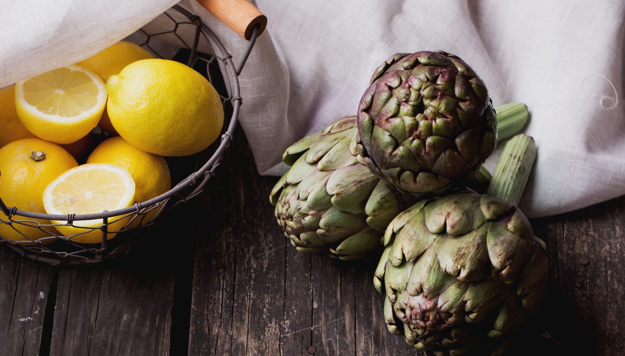 Drei Artischocken liegen auf einem dunklen Holzbrett, daneben steht ein Korb mit Zitronen Drei Artischocken liegen auf einem dunklen Holzbrett, daneben steht ein Korb mit Zitronen