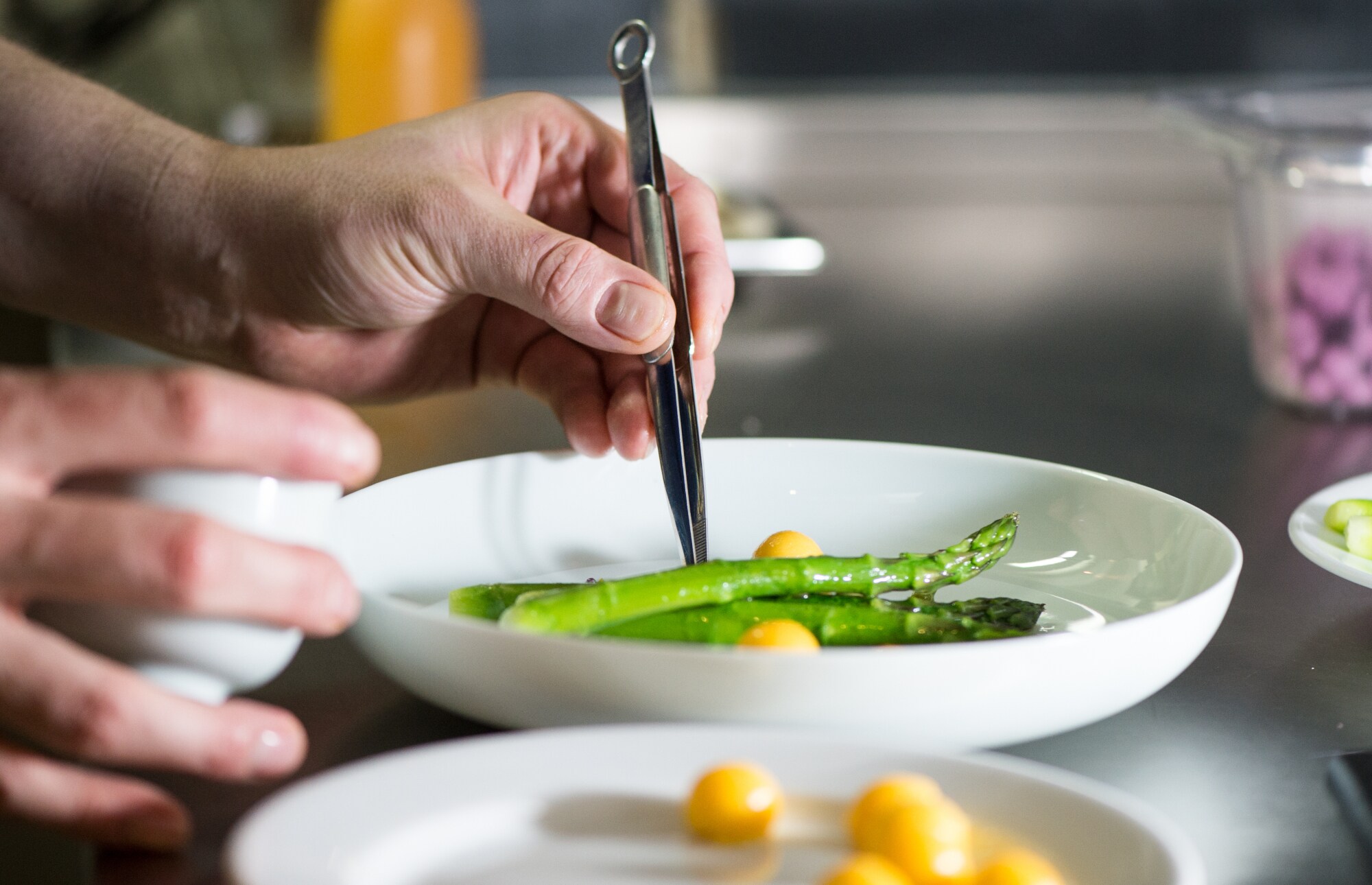 Nahaufnahme einer Hand, die mit einer Pinzette grünen Spargel und Mangostücke auf einem weißen Teller in einer Küche anrichtet