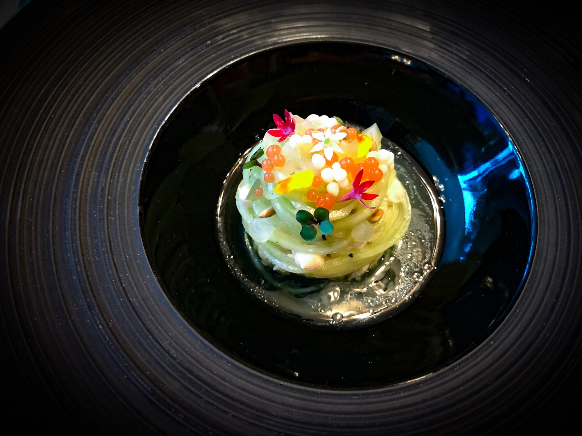 Nahaufnahme eines Gourmet-Gerichtes mit grünen, zusammengerollten Nudeln, dekoriert mit Kaviar und Blüten und angerichtet auf einem schwarzen Teller