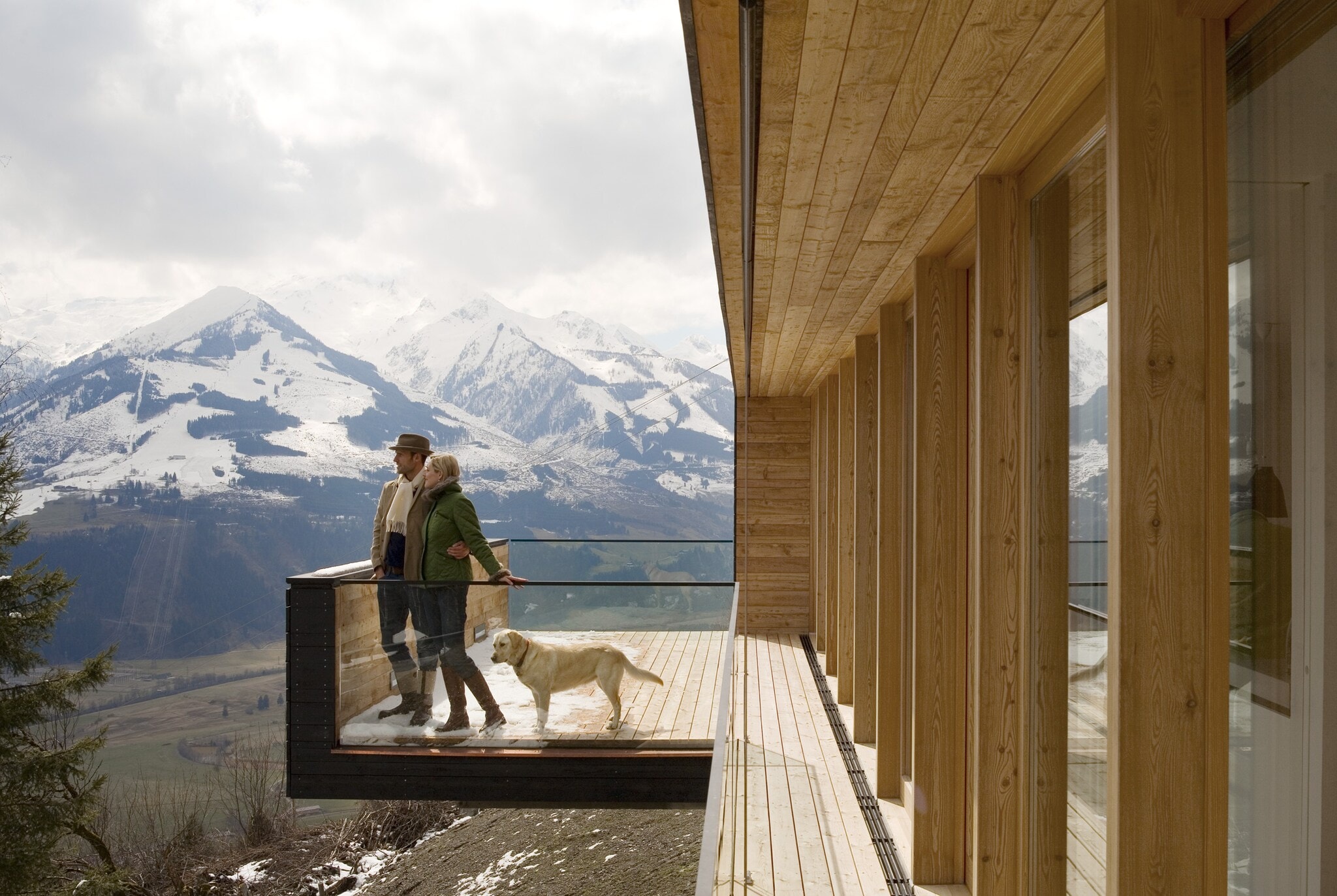 Paar mit Hund auf einer Holzterrasse eines Chalets, im Hintergrund die Berge Paar mit Hund auf einer Holzterrasse eines Chalets, im Hintergrund die Berge