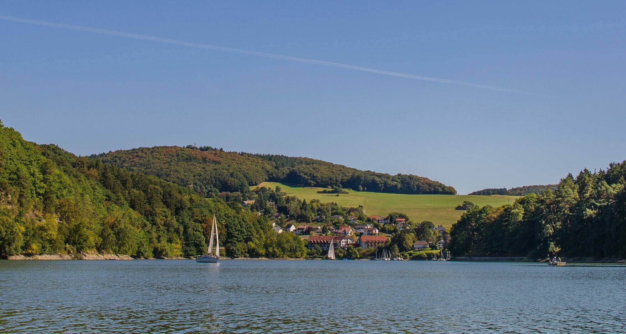 Ein See mit einem Segelschiff, dahinter hügelige Landschaft mit Wald