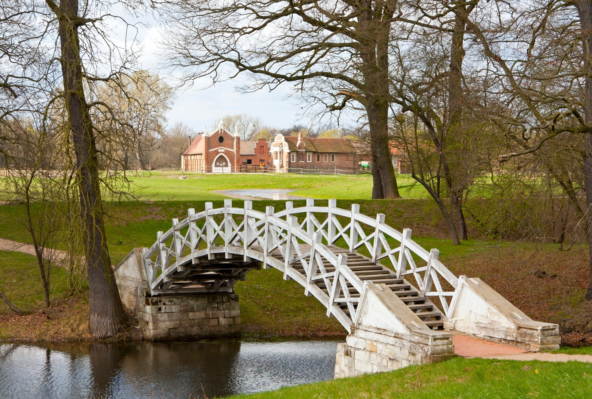 Eine kleine Brücke über einem Flusslauf, dahinter eine Wiese und ein großes Anwesen