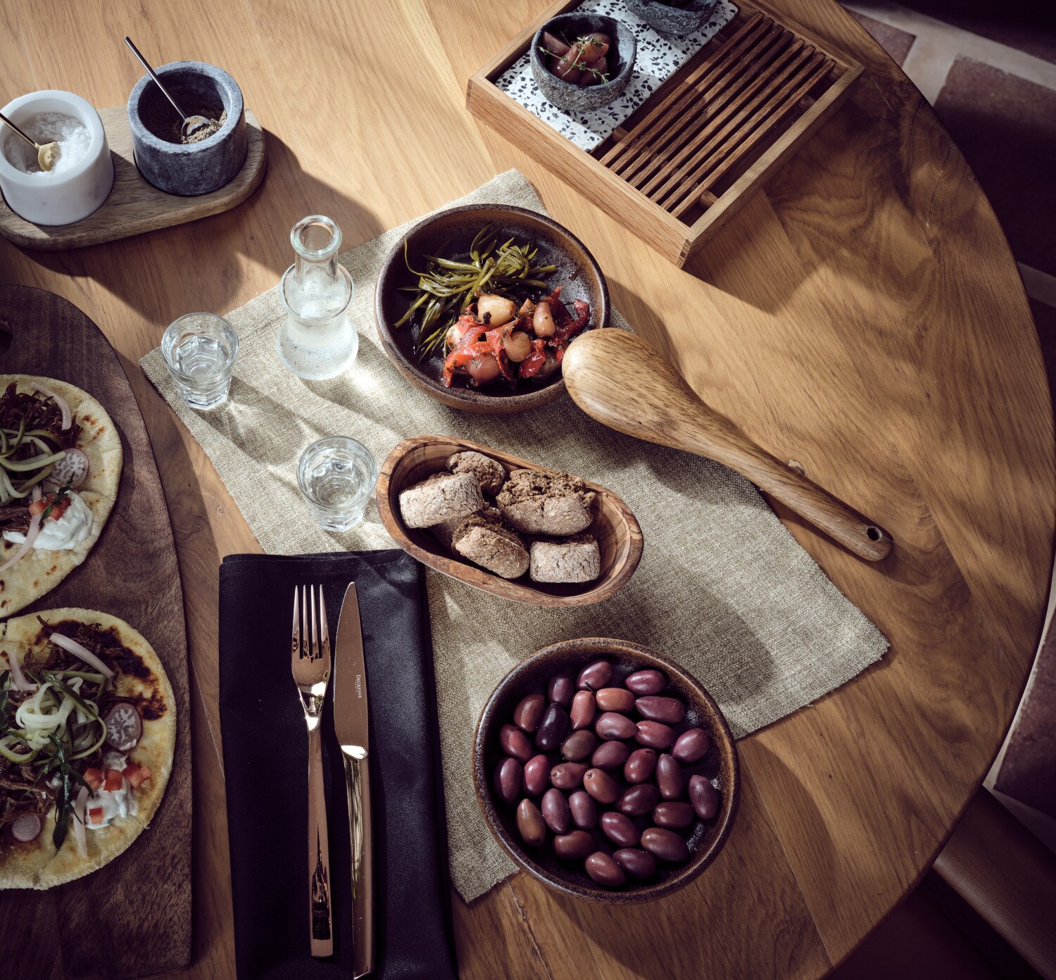 Ein gedeckter Holztisch mit Oliven, Pizzen, Brot, Gläsern, kleinen Schalen und Besteck