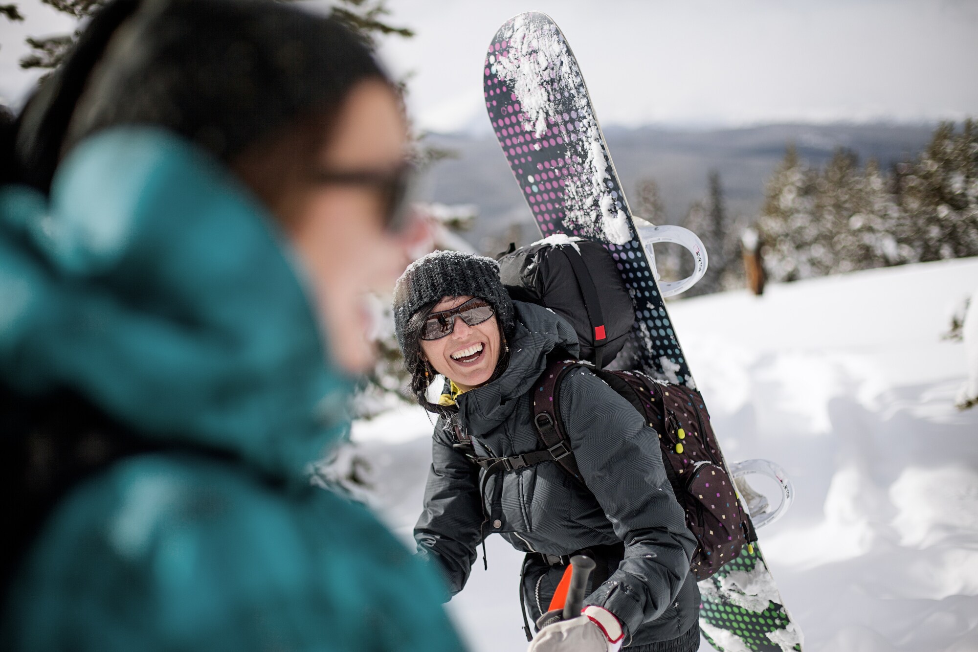 Eine lachende Frau in Winterkleidung mit einem Rucksack und einem Snowboard auf dem Rücken in einer Schneelandschaft