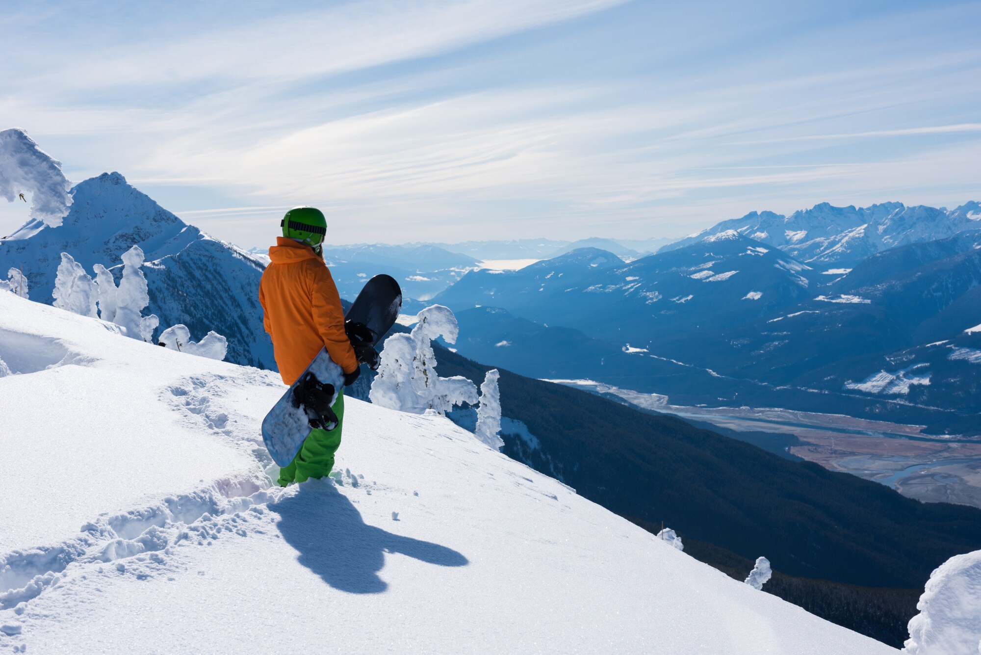 Ein Snowboarder steht mit seinem Board unterm Arm an einem Hang im Tiefschnee und blickt auf eine Berglandschaft