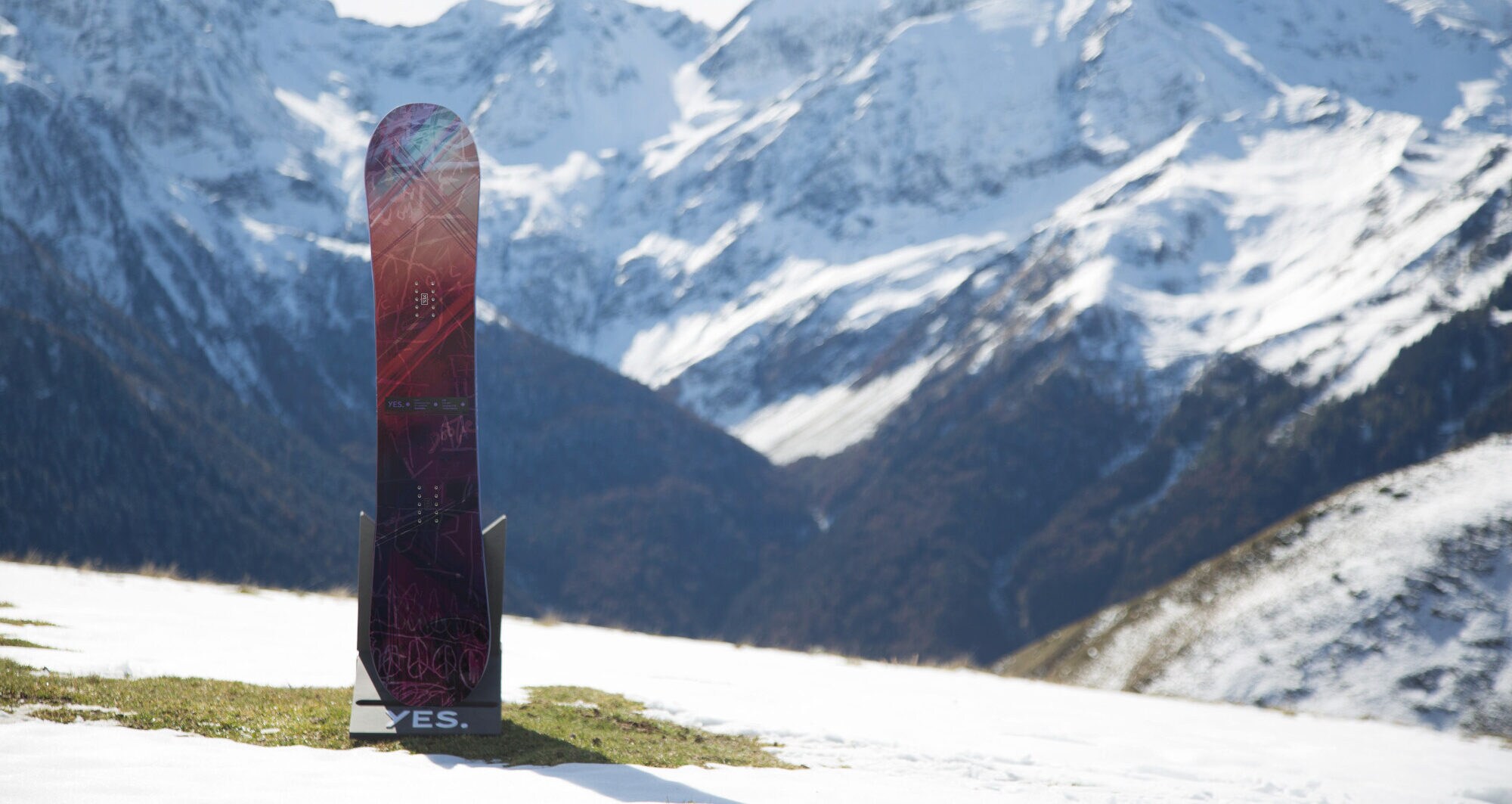 Ein Snowboard der Marke YES steht senkrecht vor einem Bergpanorma