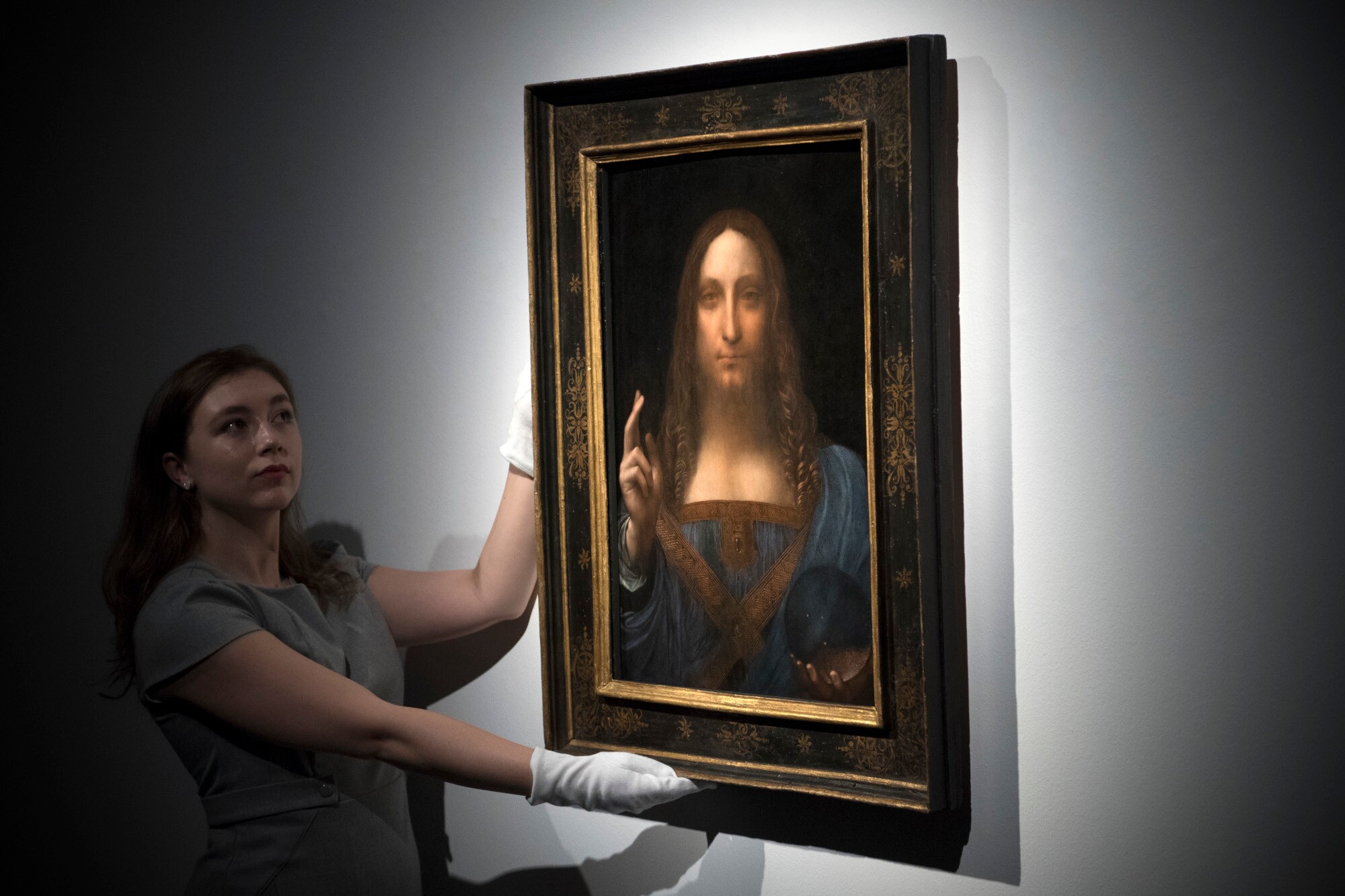 Eine Frau mit weißen Handschuhen präsentiert Leonardo da Vincis Gemälde Salvator Mundi vor einer weißen Wand