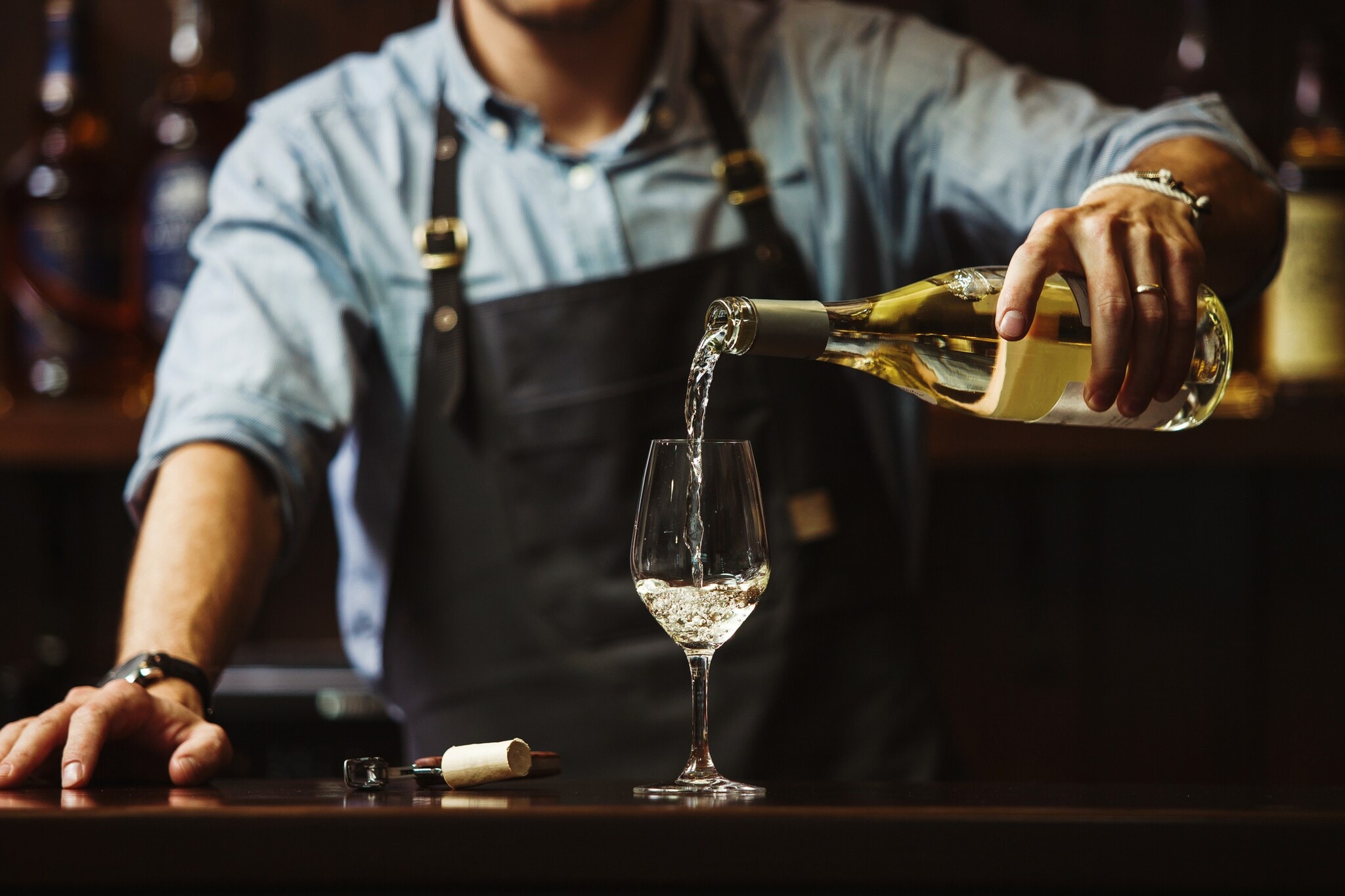 Ein Mann mit Schürze gießt Weißwein in ein Glas Ein Mann mit Schürze gießt Weißwein in ein Glas
