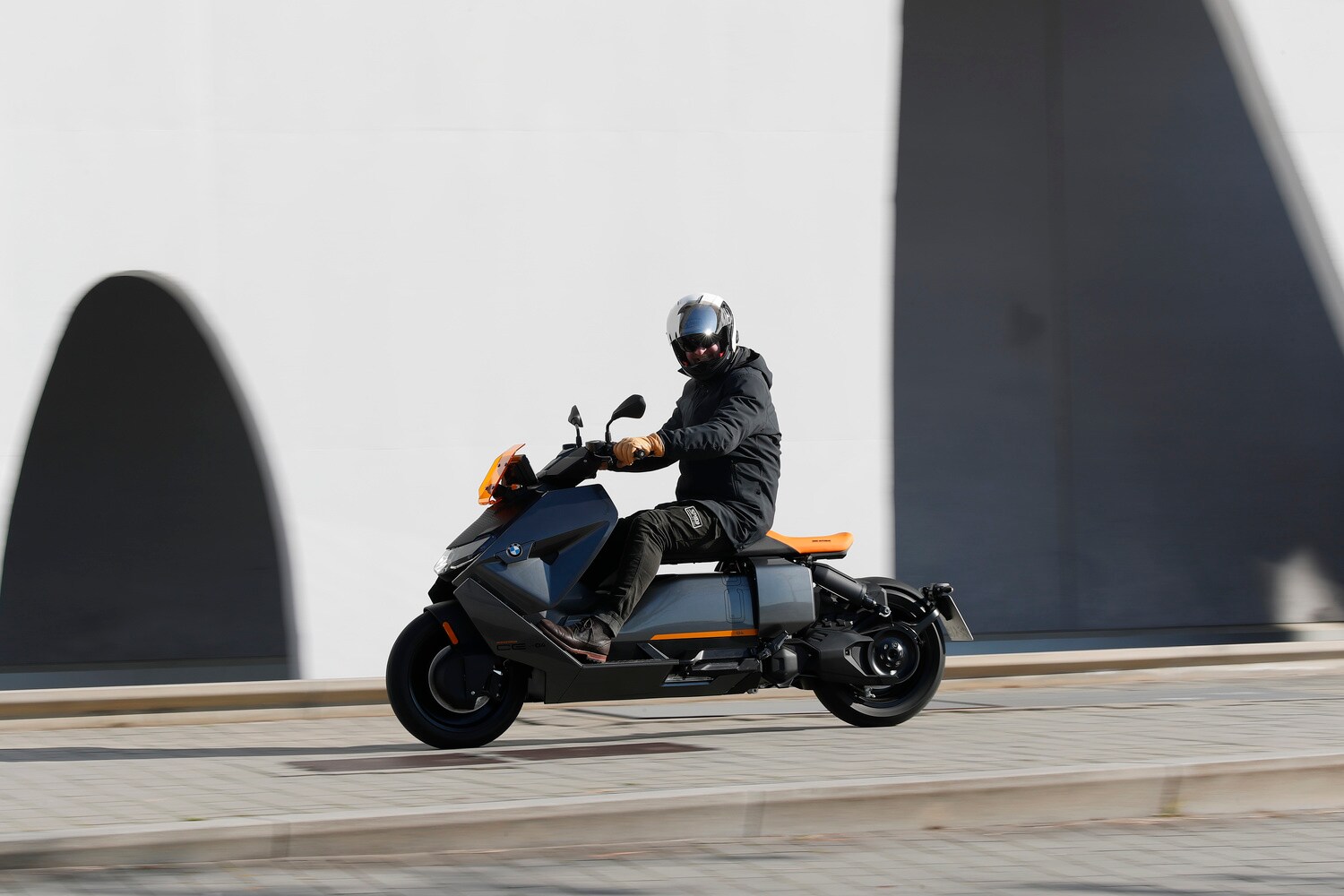 Ein grauer BMW CE 04 fährt an einer weißen Fassade vorbei, der Fahrer schaut in die Kamera. Ein grauer BMW CE 04 fährt an einer weißen Fassade vorbei, der Fahrer schaut in die Kamera.