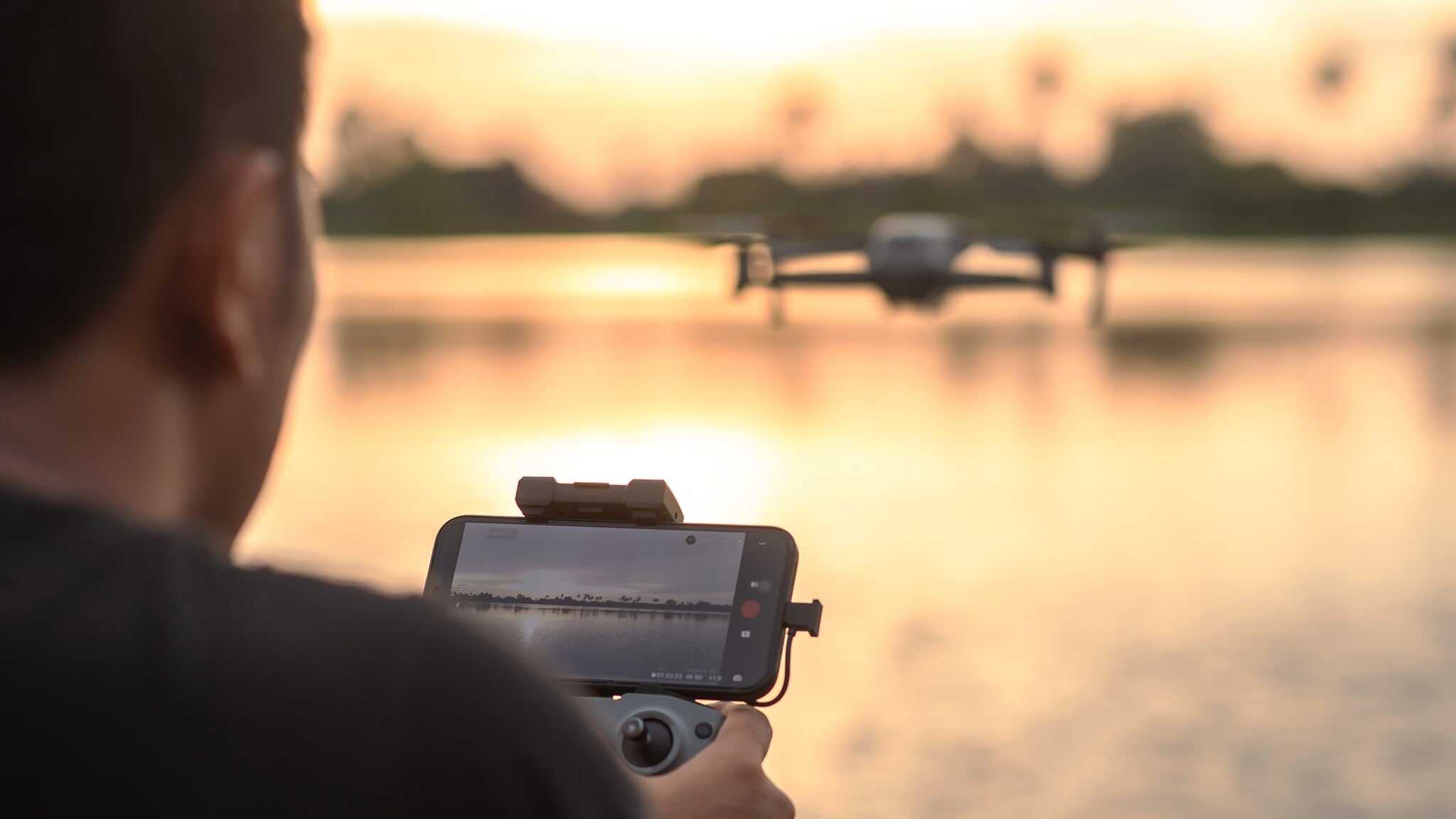 Rückansicht einer Person, die mit einer Drohne per Smartphone einen See fotografiert. Rückansicht einer Person, die mit einer Drohne per Smartphone einen See fotografiert.
