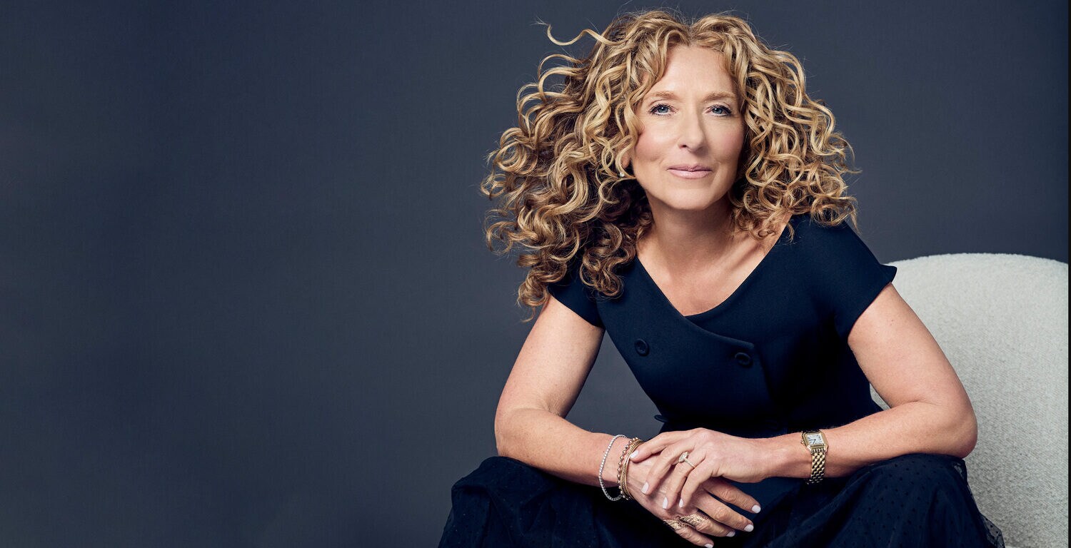 Eine Frau in blauer Kleidung mit vilden blonden Locken vor blauem Hintergrund