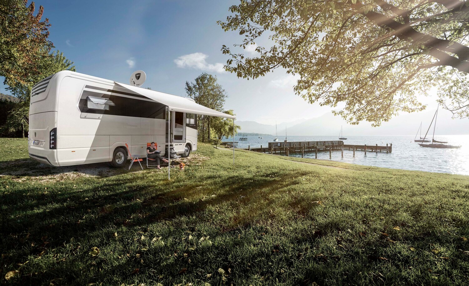 Eine Person mit Hund vor einem Luxuswohnmobil mit Blick auf einen See mit Holzsteg und Segelbooten Eine Person mit Hund vor einem Luxuswohnmobil mit Blick auf einen See mit Holzsteg und Segelbooten