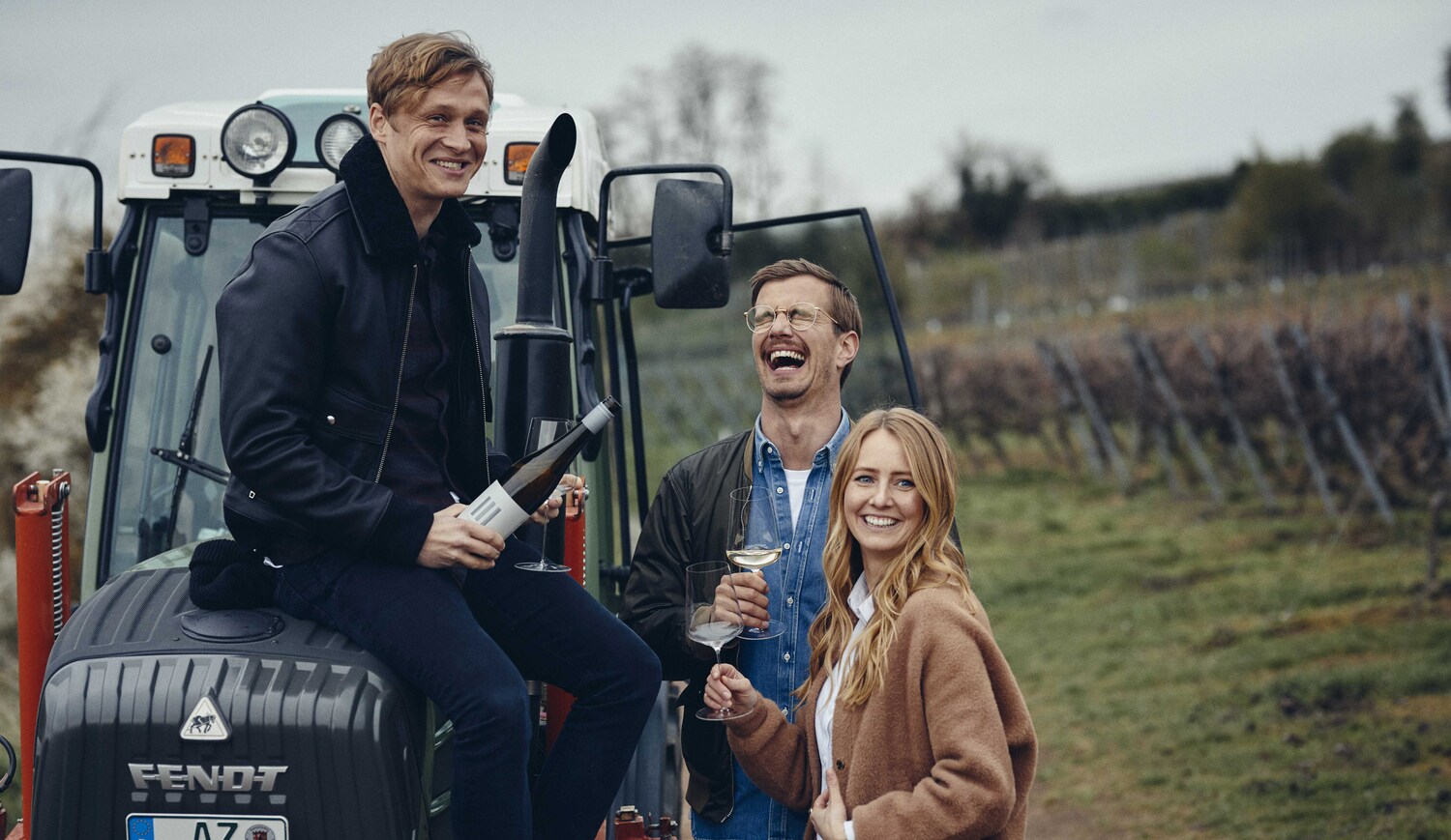 Matthias Schweighöfer sitzt lachend mit einer Weinflasche auf einem Traktor, daneben Joko Winterscheidt und Winzerin Juliane Eller auf einem Weinberg Matthias Schweighöfer sitzt lachend mit einer Weinflasche auf einem Traktor, daneben Joko Winterscheidt und Winzerin Juliane Eller auf einem Weinberg