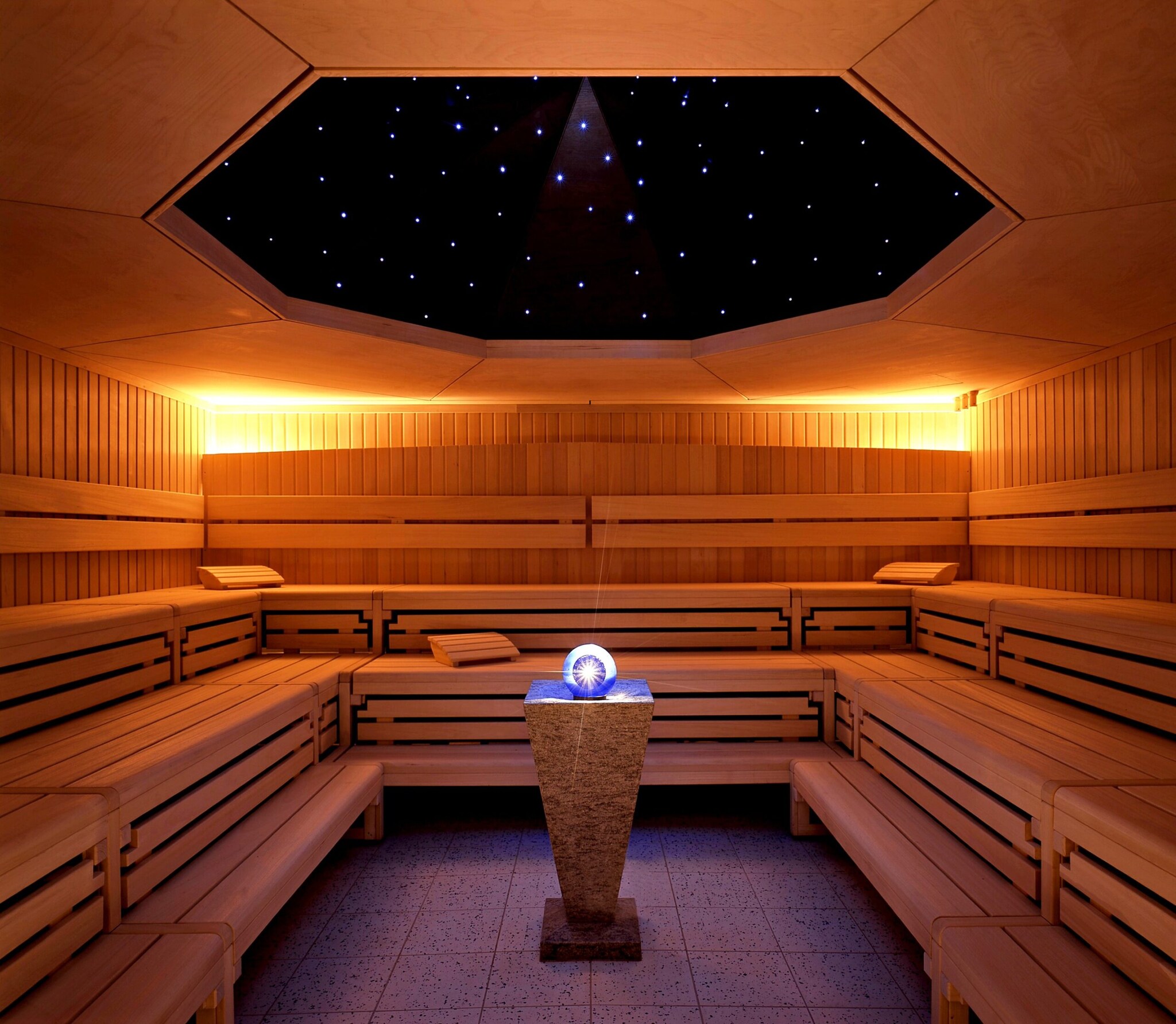 Das Innere einer Holzsauna mit Glaskugel auf einem Podest im Zentrum und einem angedeuteten, beleuchteten Sternenhimmel an der Decke