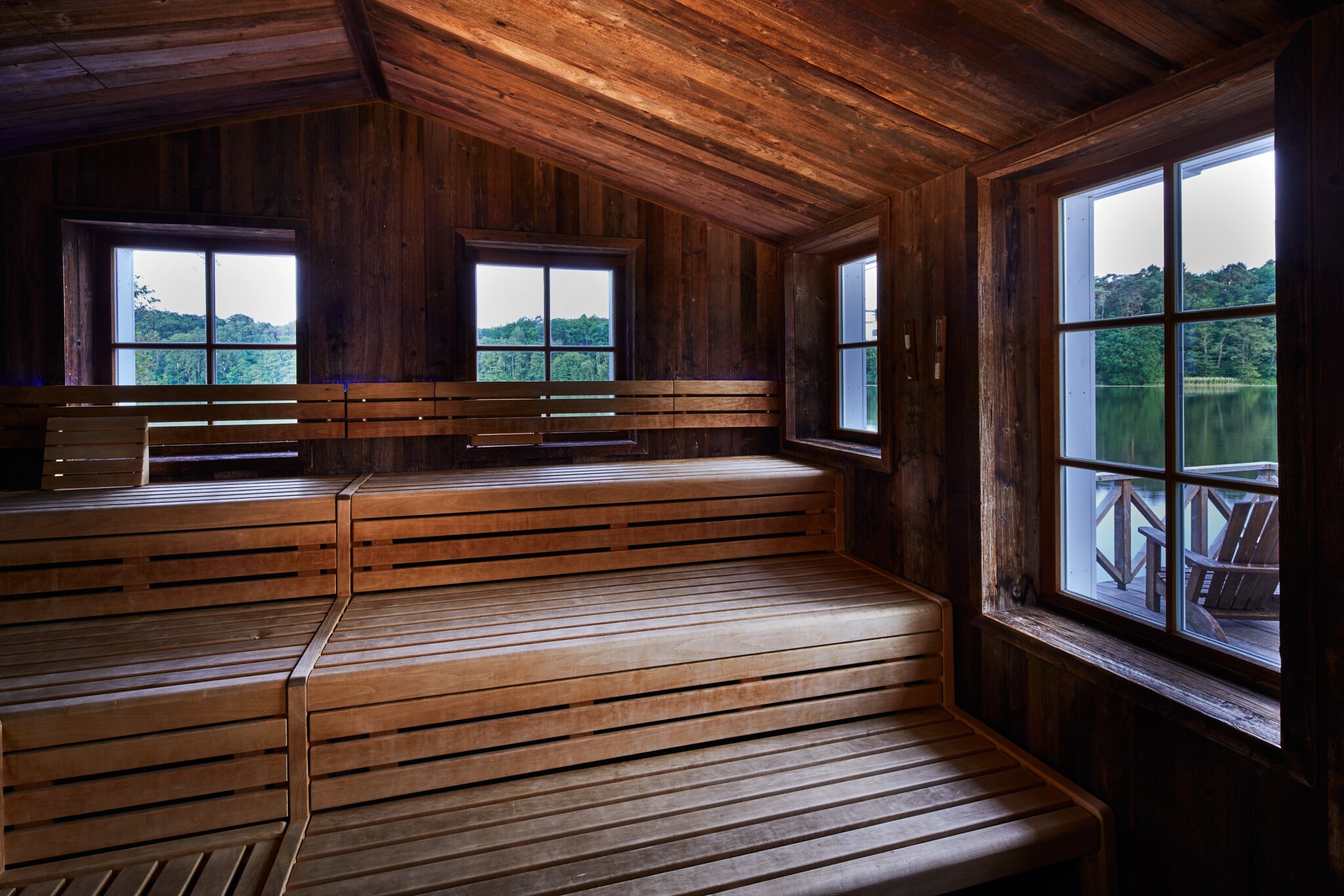 Das Innere einer Holzsauna, durch deren Fenster ein See und Waldstück zu erkennen ist Das Innere einer Holzsauna, durch deren Fenster ein See und Waldstück zu erkennen ist