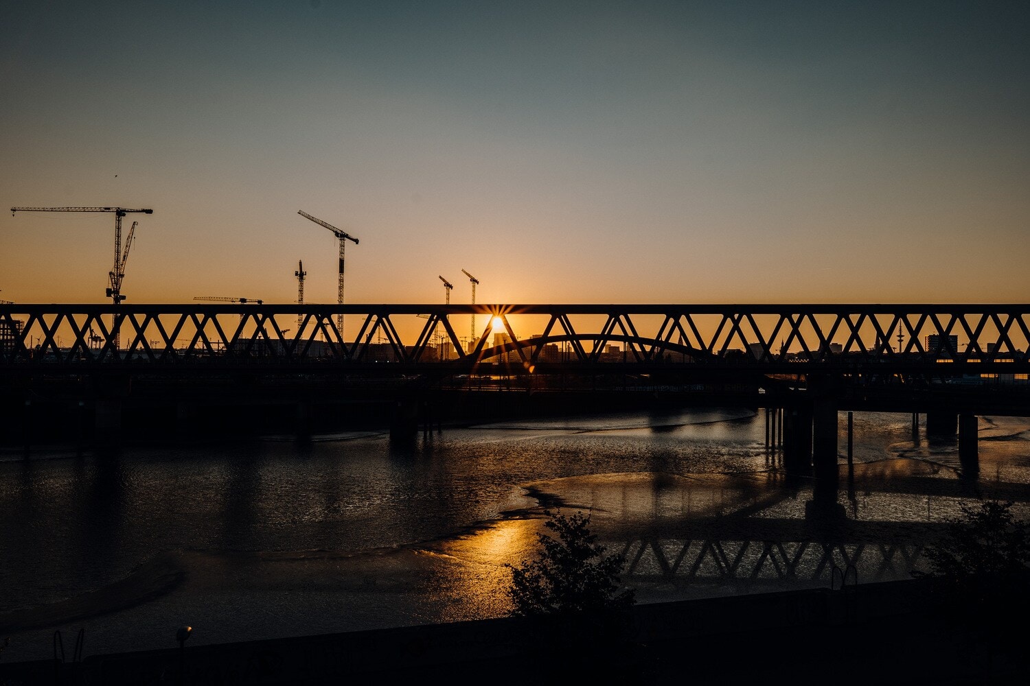 Blick auf einen Fluss, eine Eisenbrücke und Kräne bei Sonnenuntergang Blick auf einen Fluss, eine Eisenbrücke und Kräne bei Sonnenuntergang