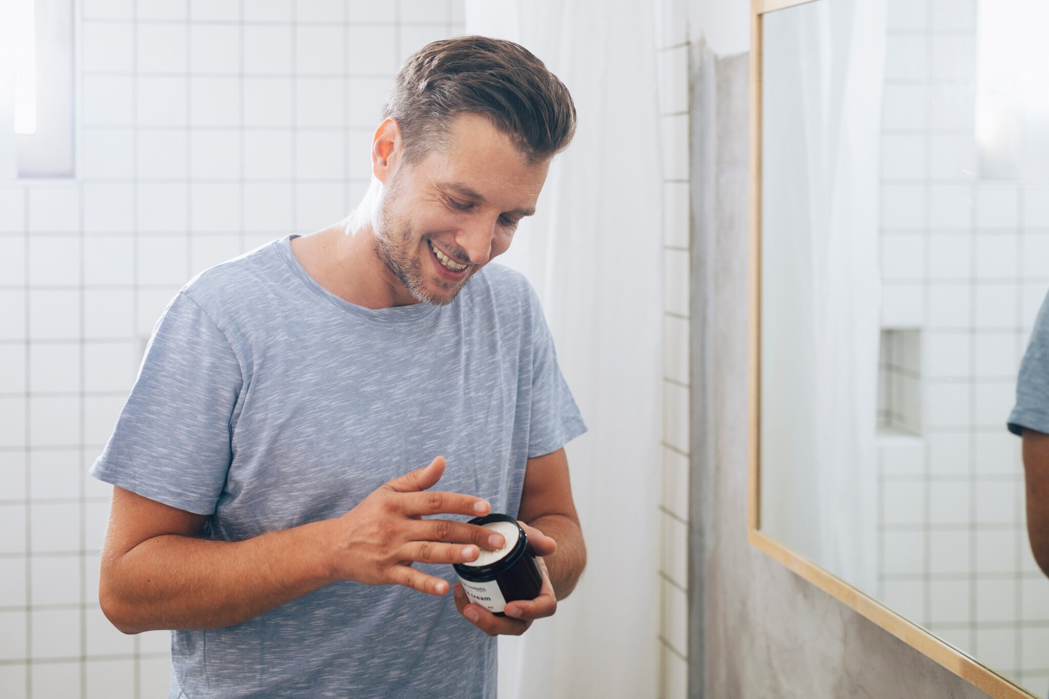 Ein lachender Mann steht im Bad und hat einen Bottich Creme in der Hand.