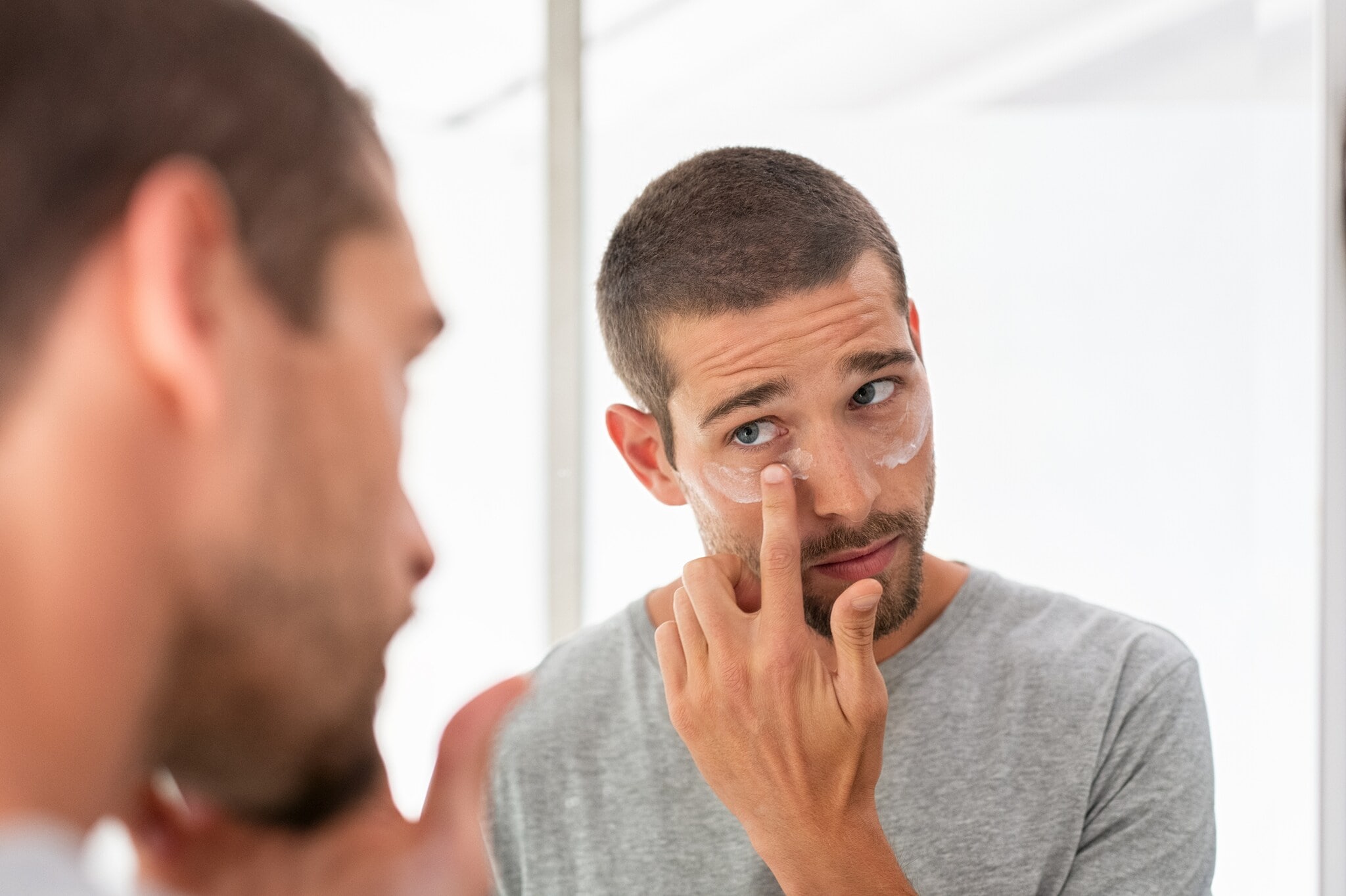 Ein Mann blickt in den Spiegel, während er sich unter den Augen Creme aufträgt Ein Mann blickt in den Spiegel, während er sich unter den Augen Creme aufträgt