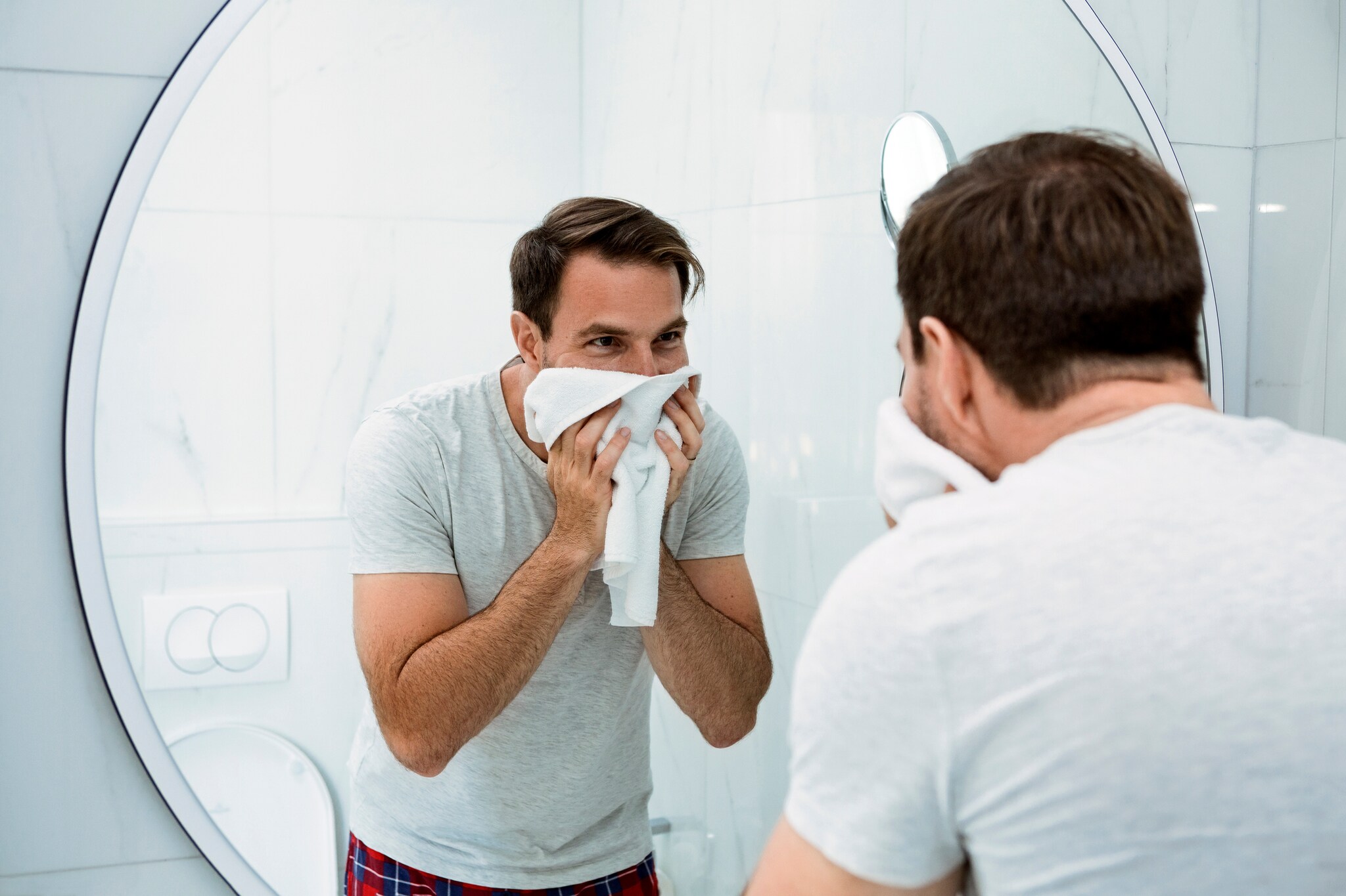 Ein Mann trocknet sich im Badezimmer sein Gesicht mit einem Handtuch ab und blickt dabei in den Spiegel.