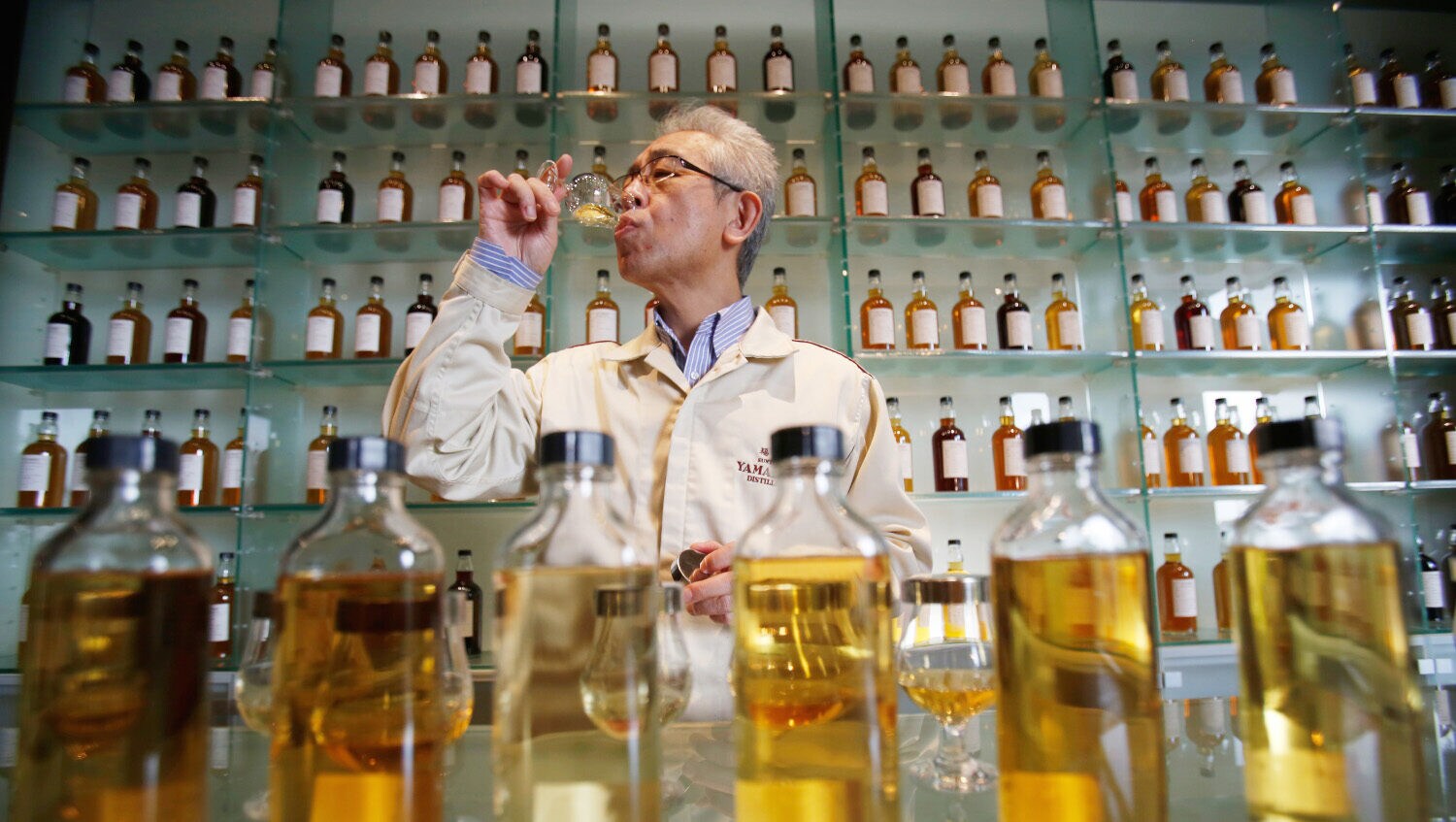 Ein Mann in einem weißen Kittel verkostet Whisky in einer Destillerie vor einem Regal mit Flaschen