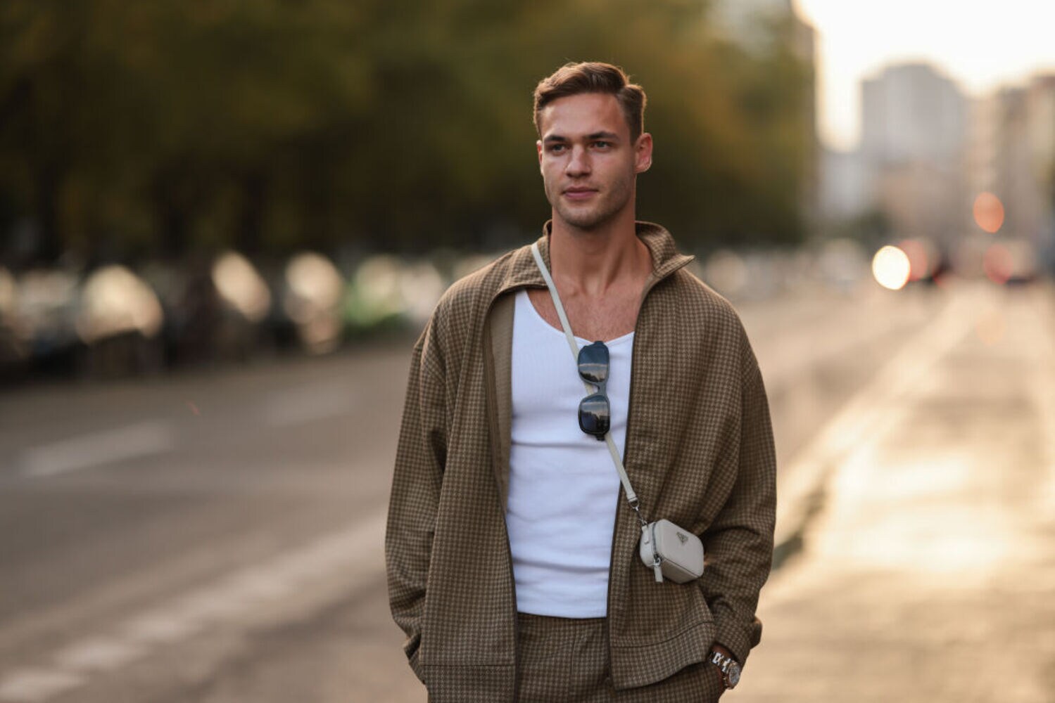 Ein Mann trägt einen braunen Trainingsanzug, hat eine Sonnenbrille am Shirt und eine kleine Tasche umgehängt.