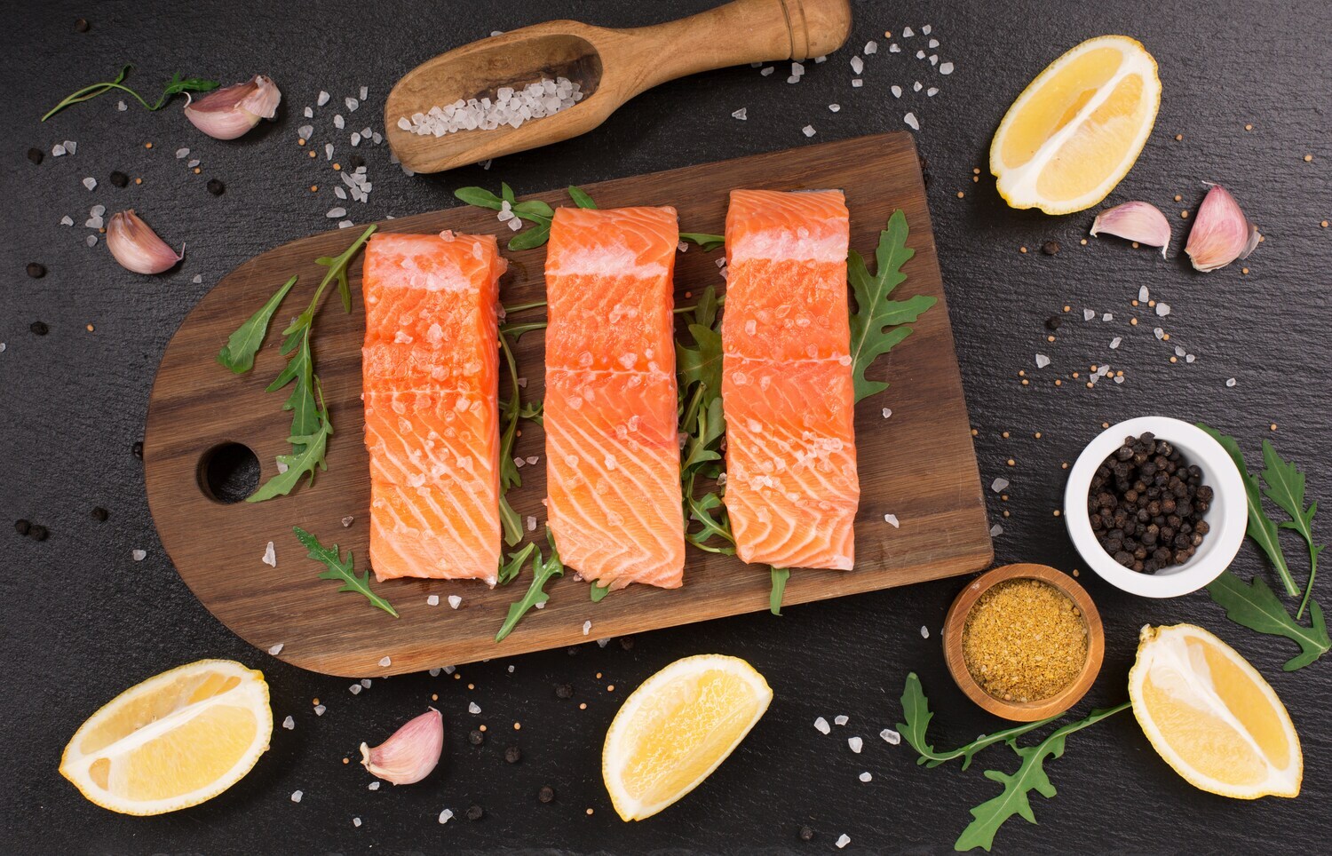 Drei Lachsfilets auf einem Holzbrett, dekoriert mit Zitrone, Knoblauch und Gewürzen.