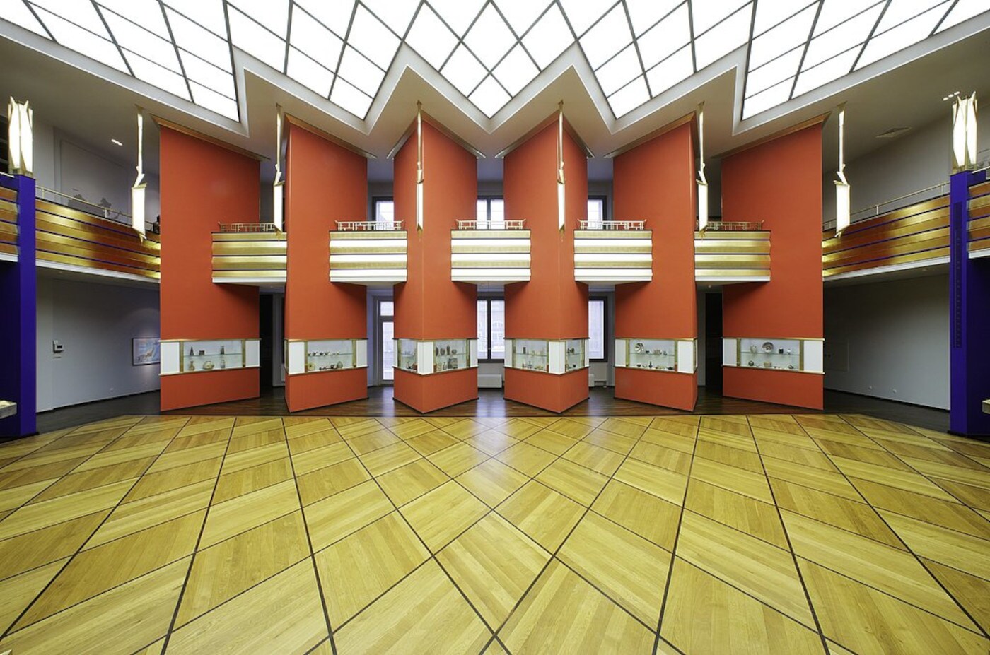 Ein geometrisch angelegter Raum mit Holzfußboden, orangen Säulen und einer Empore