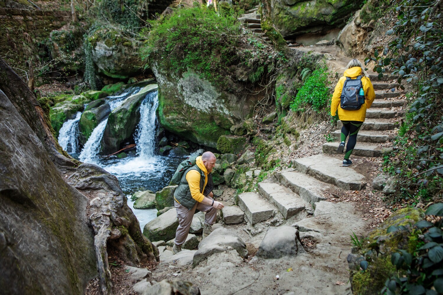Zwei Wanderer steigen neben einem kleinen Wasserfall Steinstufen hinauf. Zwei Wanderer steigen neben einem kleinen Wasserfall Steinstufen hinauf.