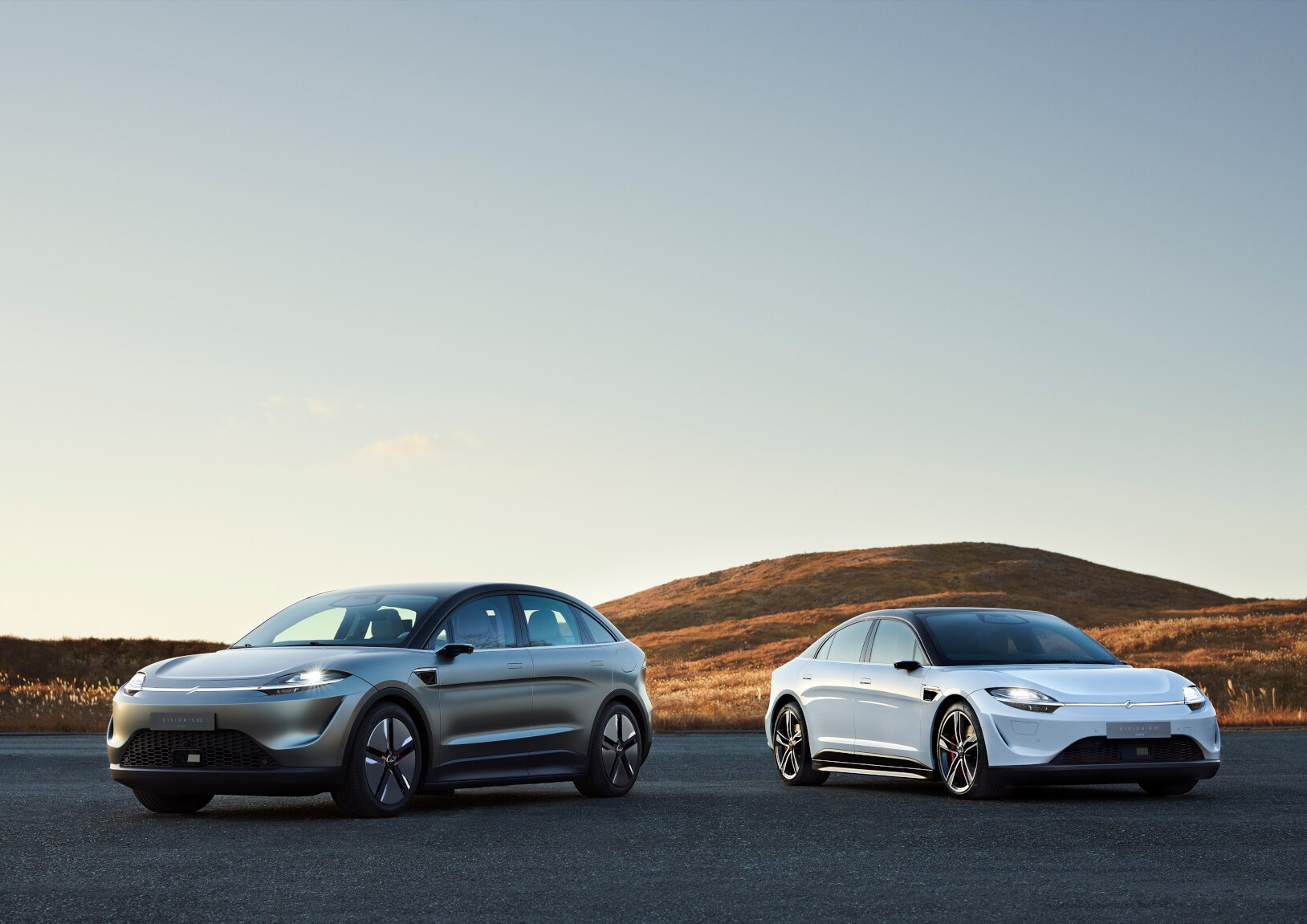 Ein weißes und ein silbernes Auto auf einem Parkplatz, im Hintergrund ein Hügel