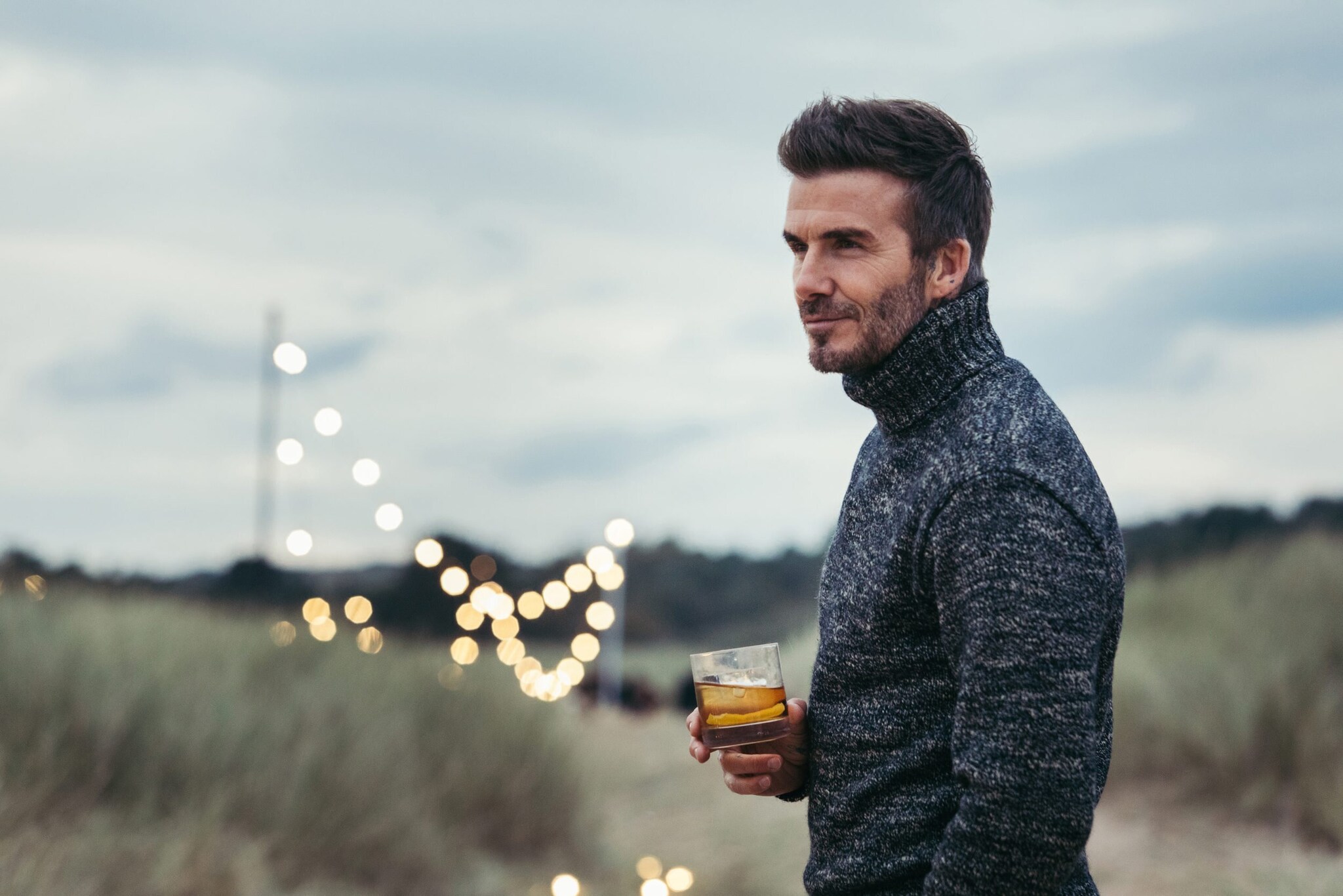 Porträtaufnahme von David Beckham in den Dünen mit einem Glas Whisky in seiner Hand