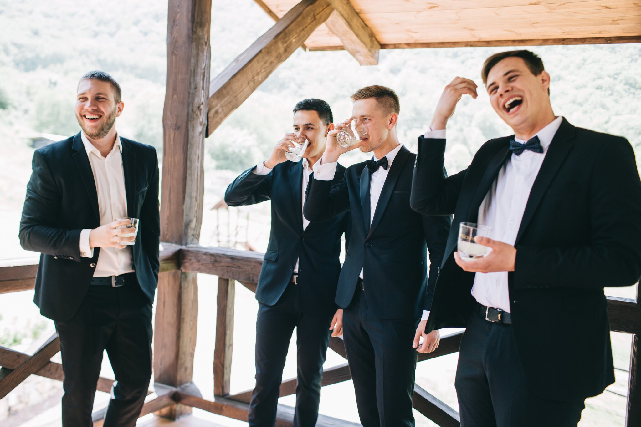 Vier Männer in Anzügen, teilweise mit Fliege, stehen lachend unter einem Holzdach