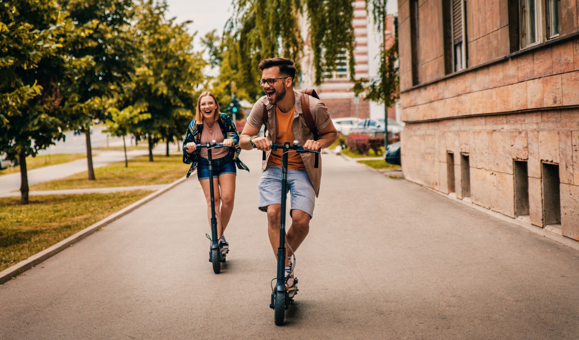 Ein junges Paar fährt lachend auf E-Scootern über einen breiten Gehweg Ein junges Paar fährt lachend auf E-Scootern über einen breiten Gehweg