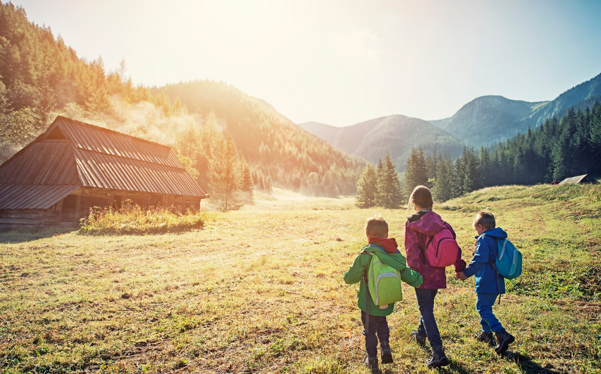 Drei Kinder laufen Hand in Hand in einem bergigen Gebiet auf eine Holzhütte zu
