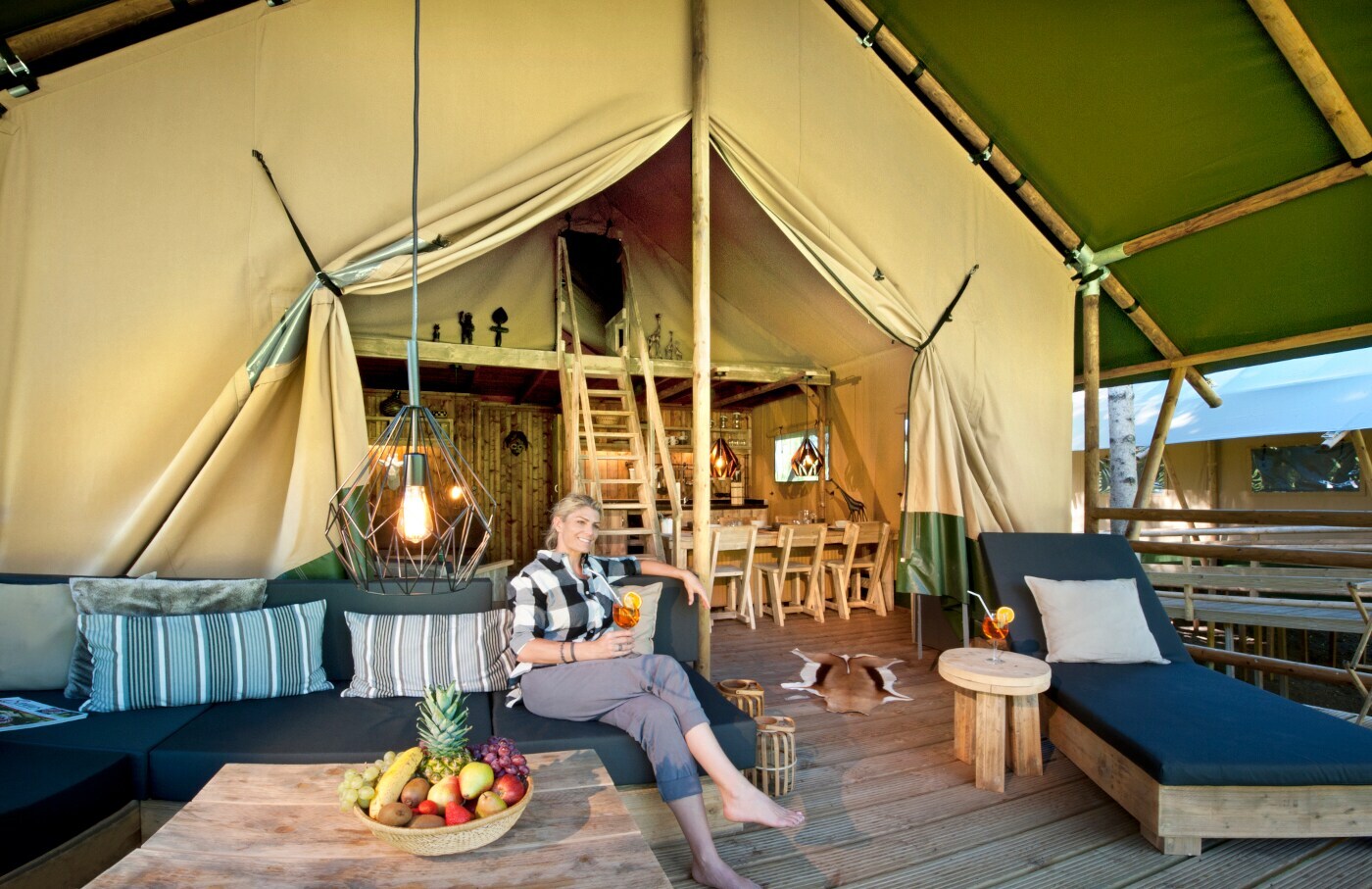 Eine Frau sitzt vor einem luxuriös ausgestatteten Zelt mit einem Cocktail in der Hand auf einem Sofa