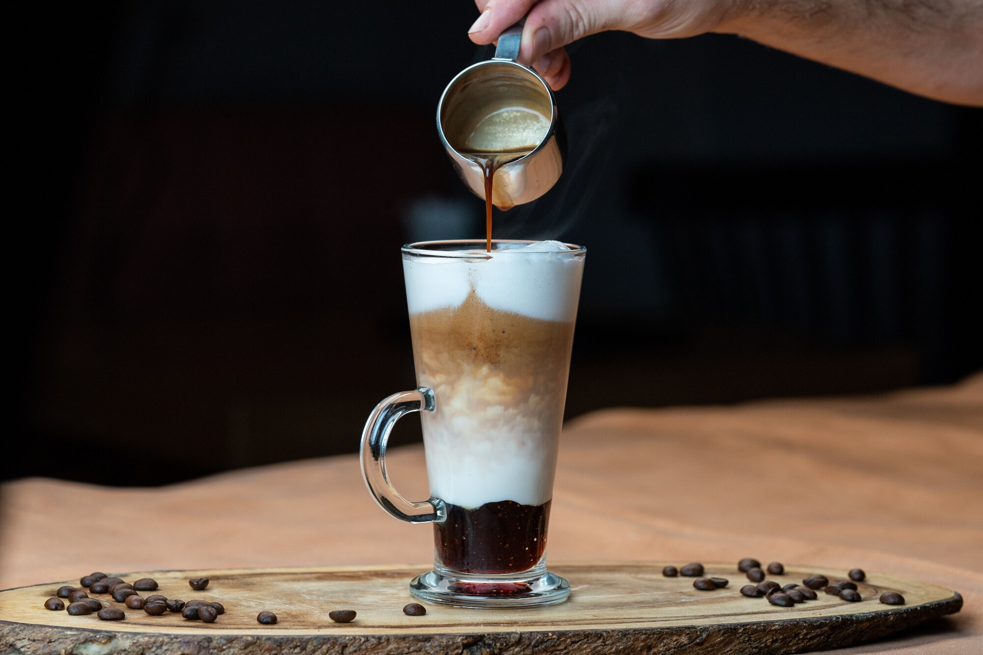 Espresso wird aus einem Kännchen in ein Glas mit Milchschaum gegossen Espresso wird aus einem Kännchen in ein Glas mit Milchschaum gegossen