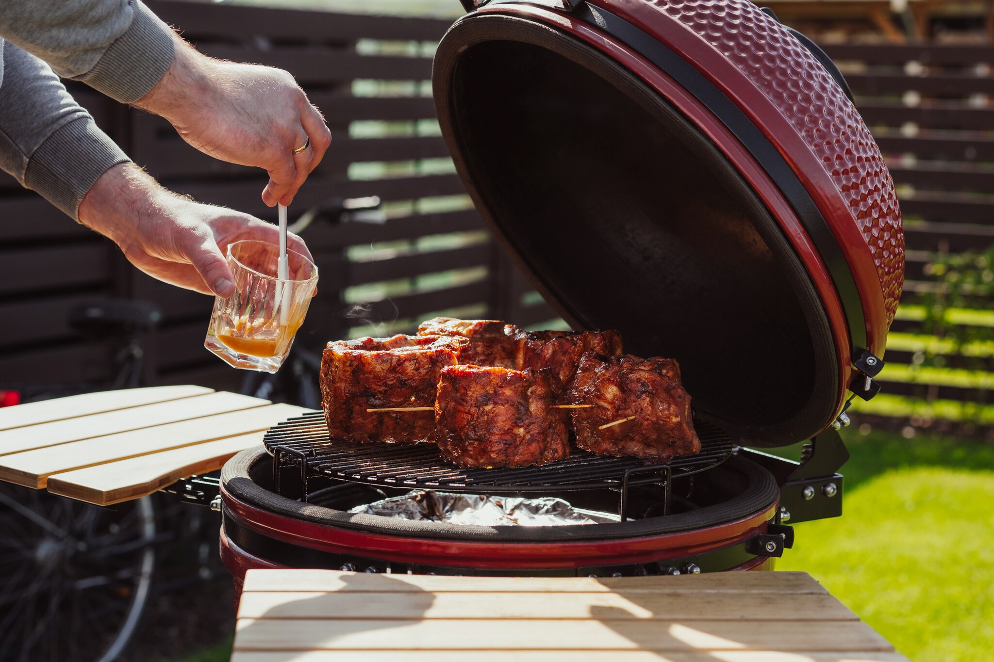 Roter Keramikgrill mit Rippchen auf dem Rost, die gerade von einem Mann mit Marinade bestrichen werden