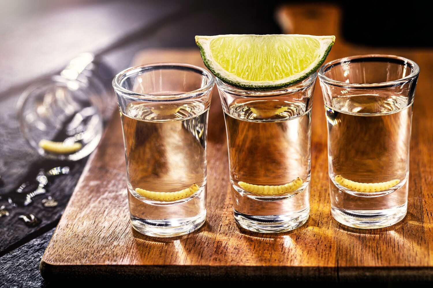 Drei Tequila-Shots auf einem Holzbrett, garniert mit Limette und Wurm