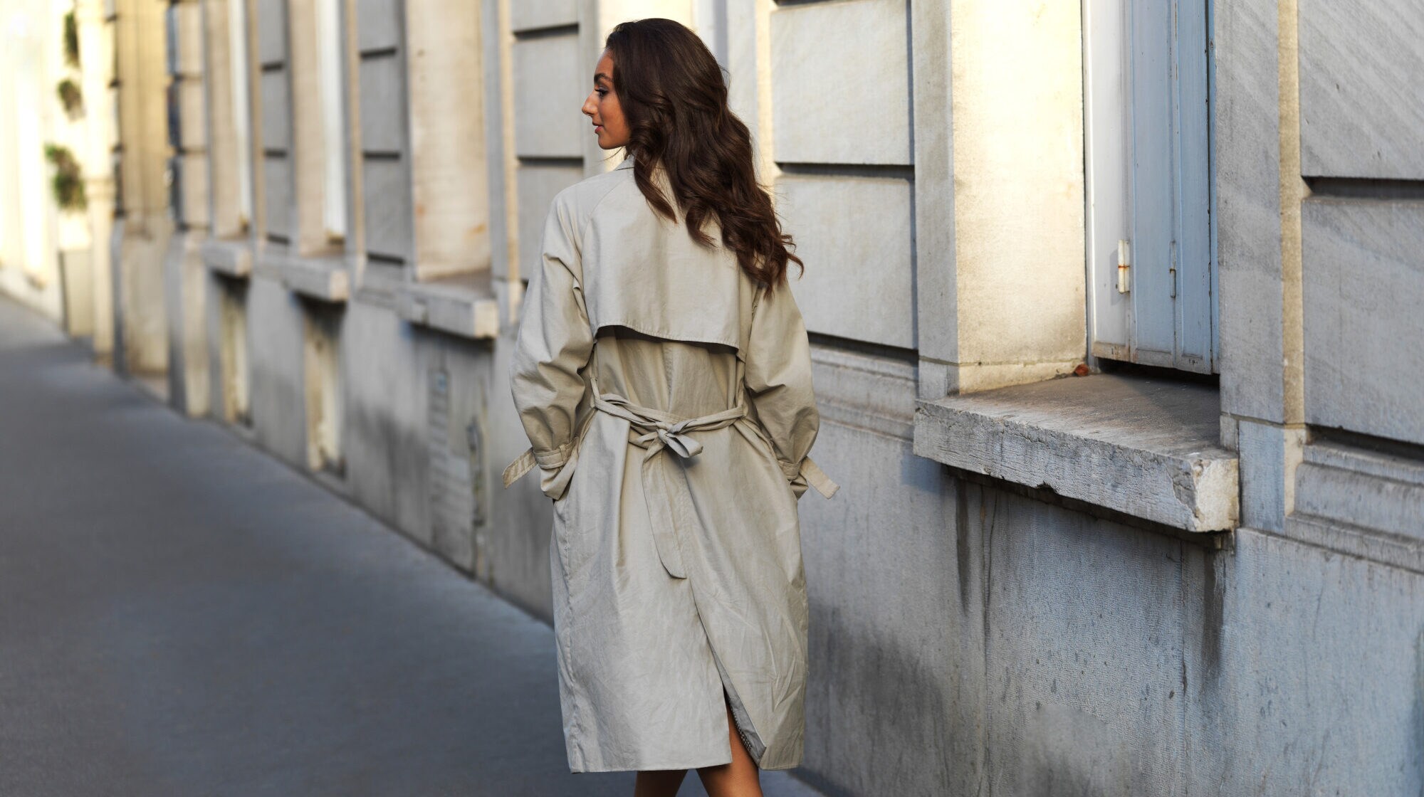 Rückansicht einer gehenden Frau im beigen Trenchcoat auf einer Straße