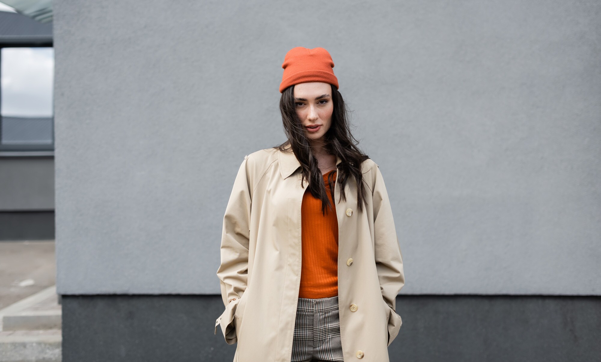 Eine stylishe, junge Frau, gekleidet in einen beigen Trenchcoat zu orangefarbenem Pullover und Mütze, steht lässig vor einer Betonwand