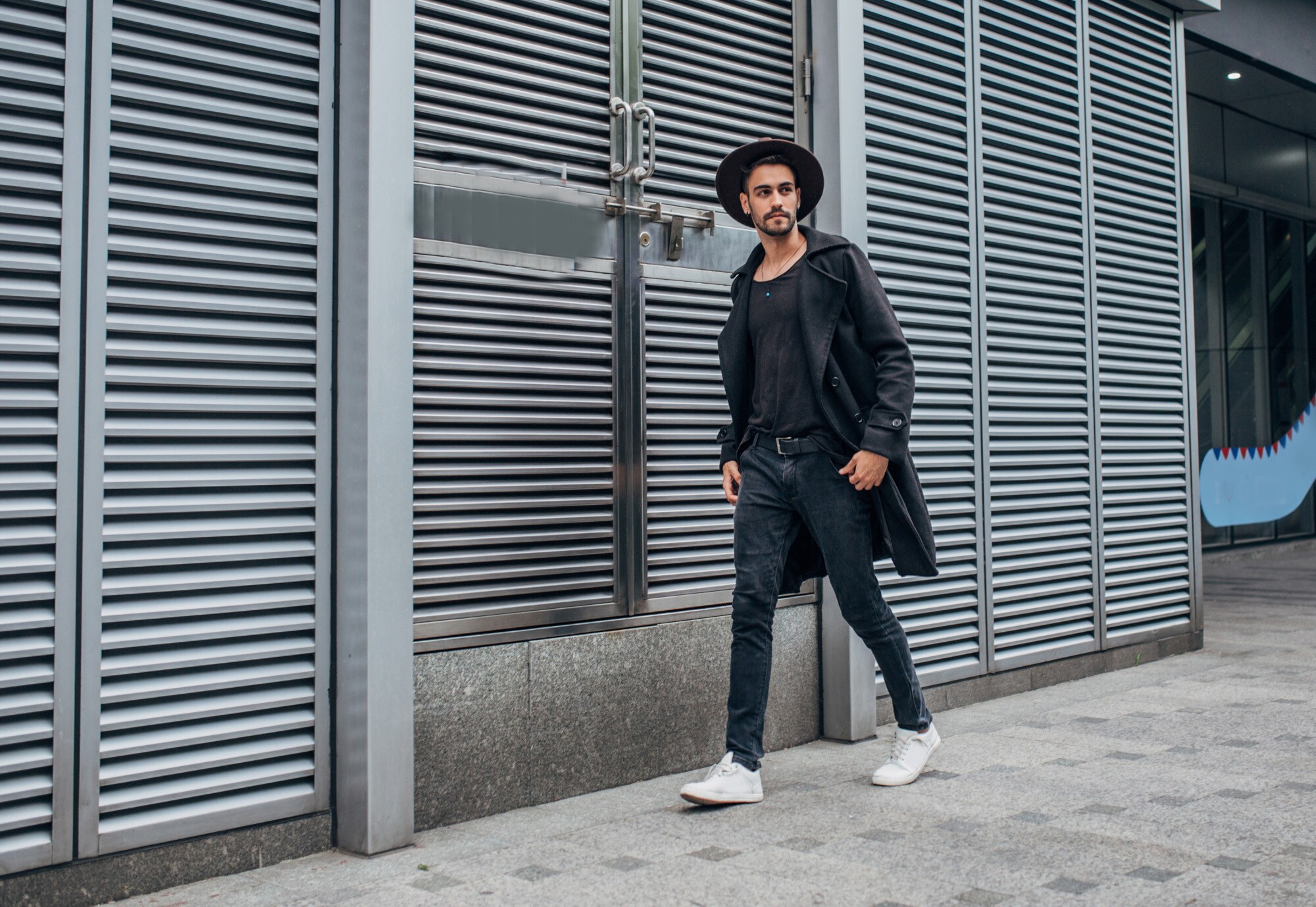 Ein stylisher Mann mit Hut und schwarzem Trenchcoat läuft lässig auf einem Gehweg