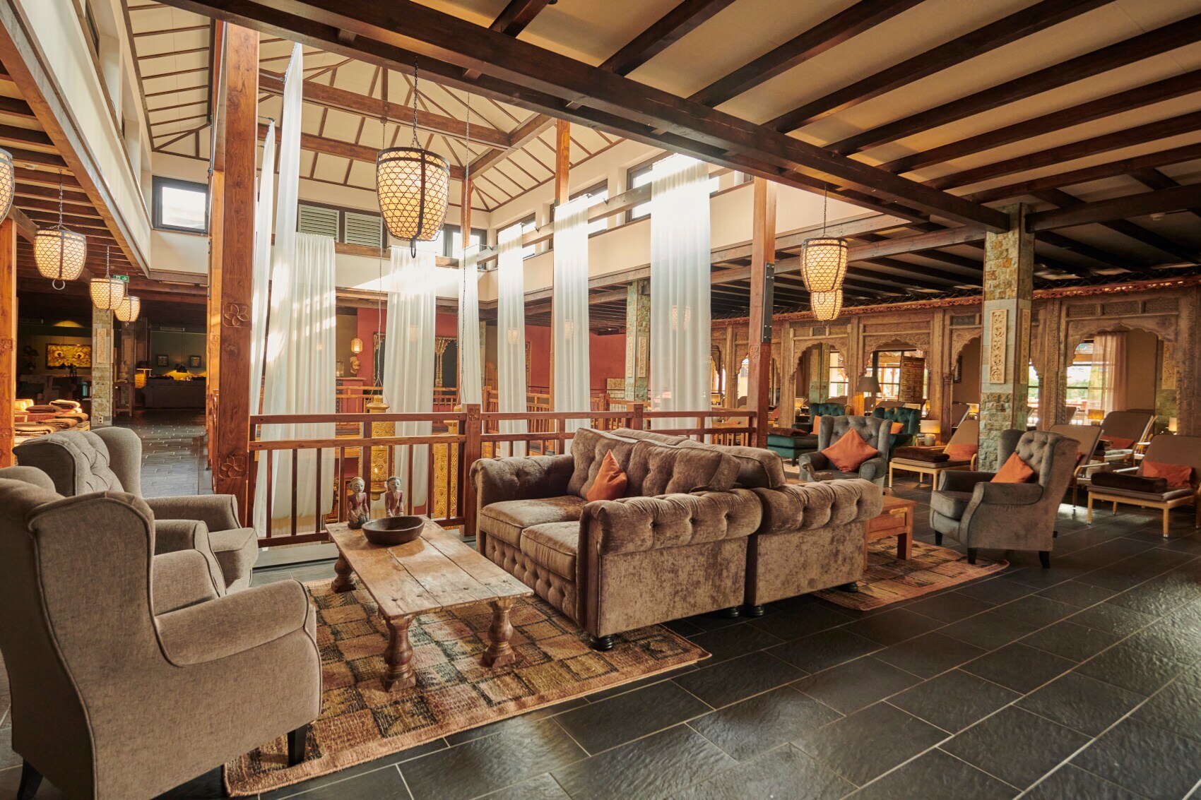 Der Innenbereich im vabali spa mit grauen Sitzgelegenheiten, weiß-transparenten Vorhängen, bunten Teppichen und einem Holzgeländer.