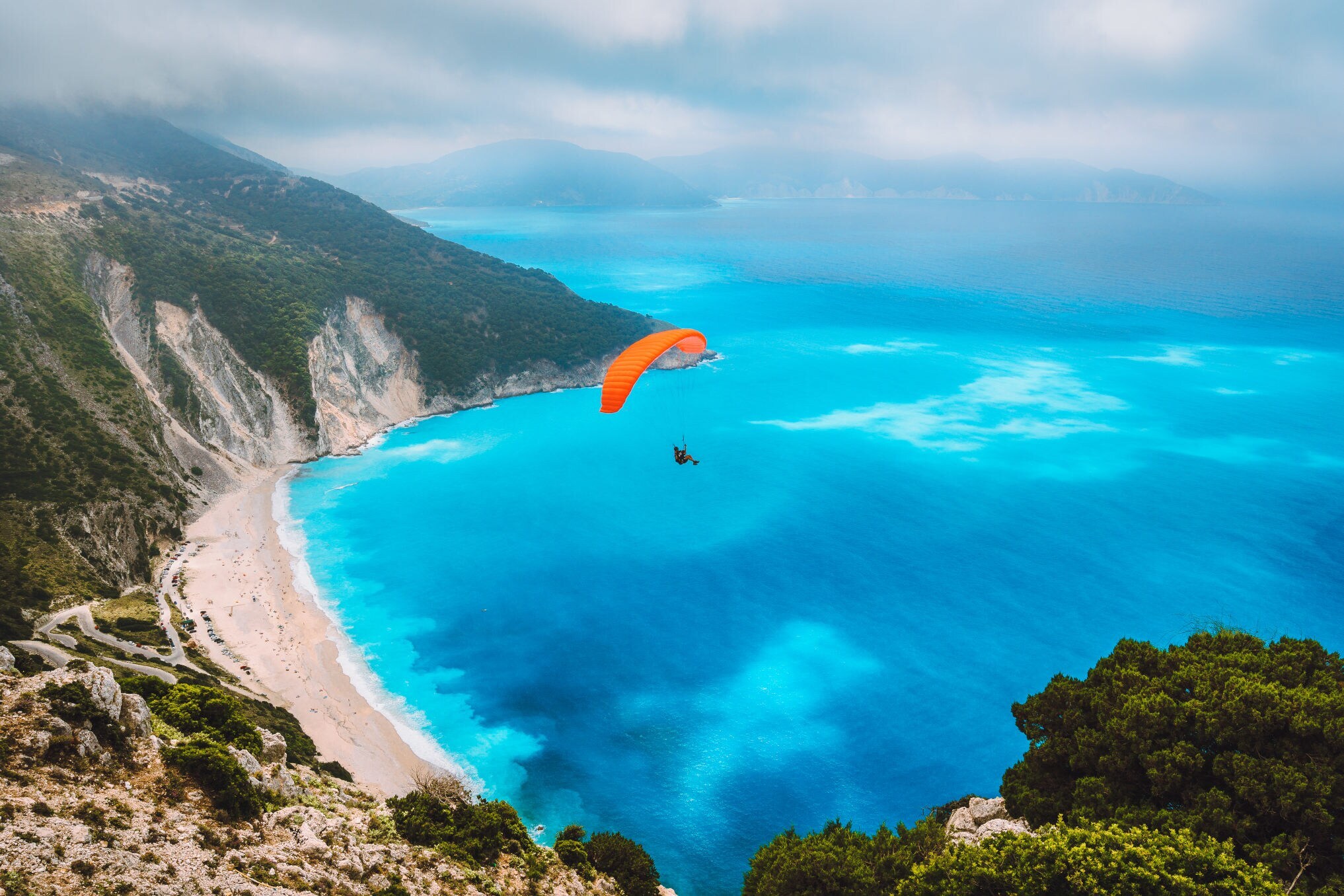 Ein Gleitschirmflieger über einer Badebucht an einer Bergküste mit Sandstrand und türkisblauem Wasser.
