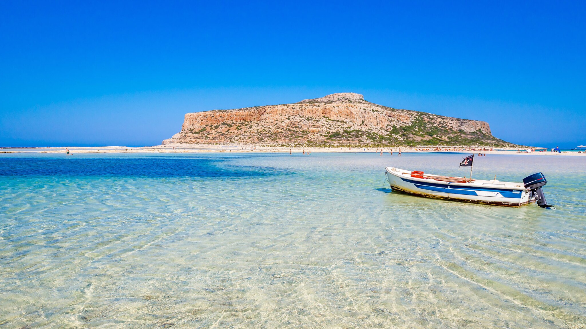 Lagunenstrand von Balos mit Blick auf türkisblaues Wasser, Motorboot und kleiner Insel am Horizont.